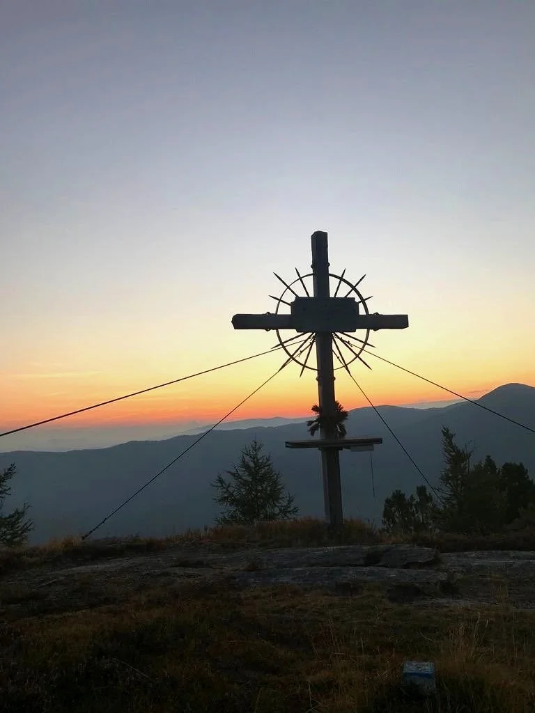 Gipfelkreuz auf einem Berg bei Sonnenaufgang, umgeben von Bäumen, mit einem klaren Himmel und Bergen im Hintergrund.