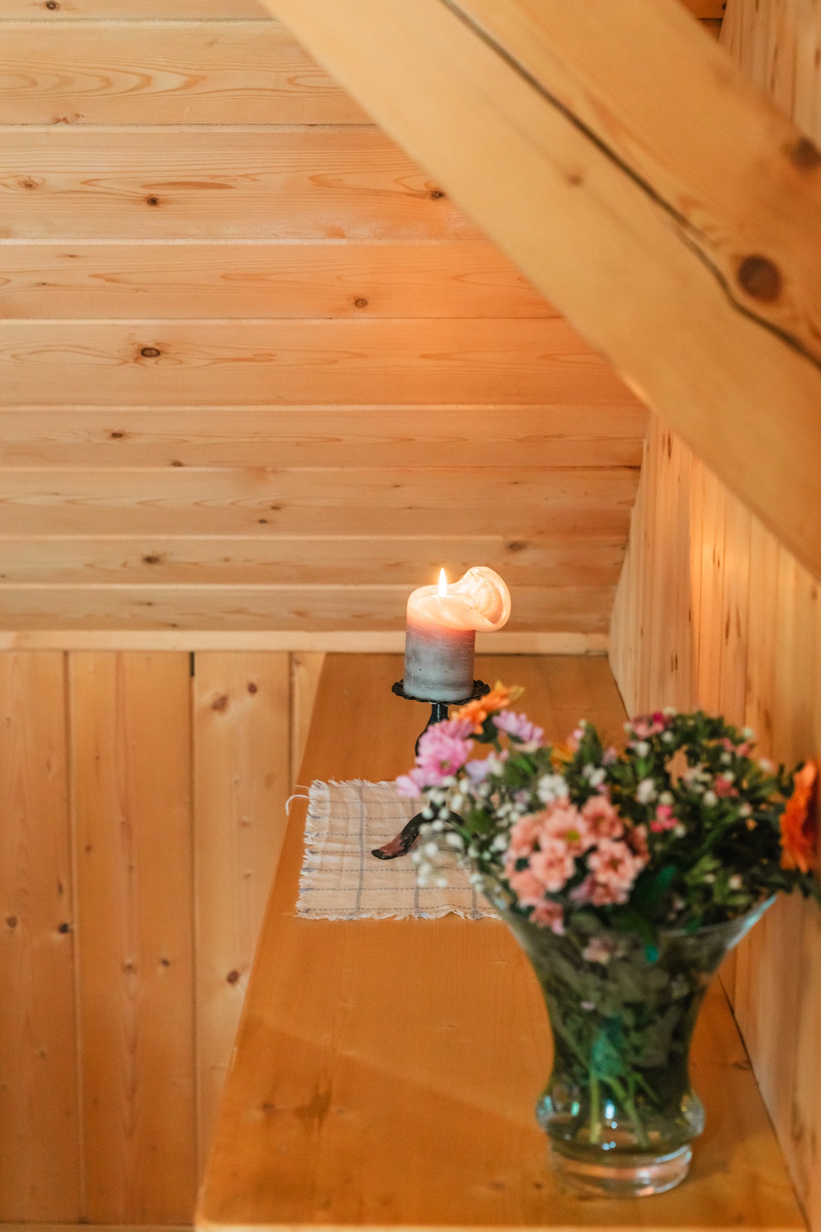 Ein Holzregal in einem Raum mit Holzwänden. Auf dem Regal stehen eine brennende Kerze und ein Strauß frischer Blumen in einer Glasvase. Ein Tuch liegt unter der Kerze.