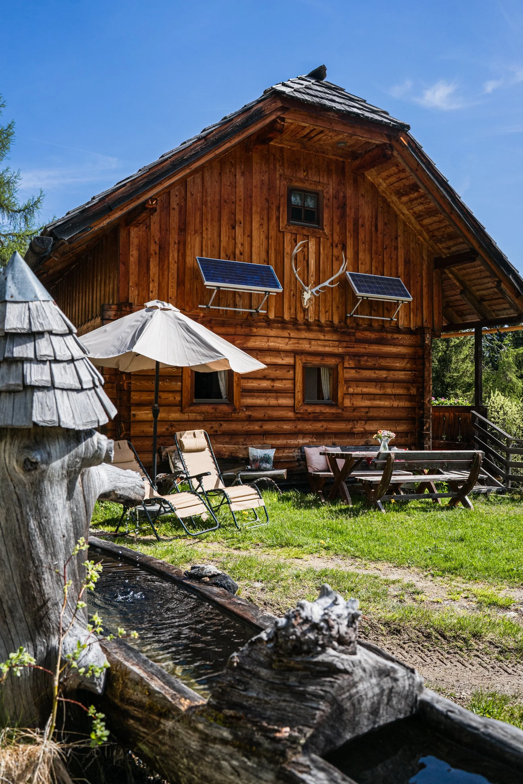 Eine Almhütte mit Solarpaneelen an der Vorderseite, Liegestühle, ein Sonnenschirm und ein Holztisch mit Bänken im Garten, sowie ein rustikaler Holzbrunnen im Vordergrund, umgeben von grüner Vegetation unter blauem Himmel.