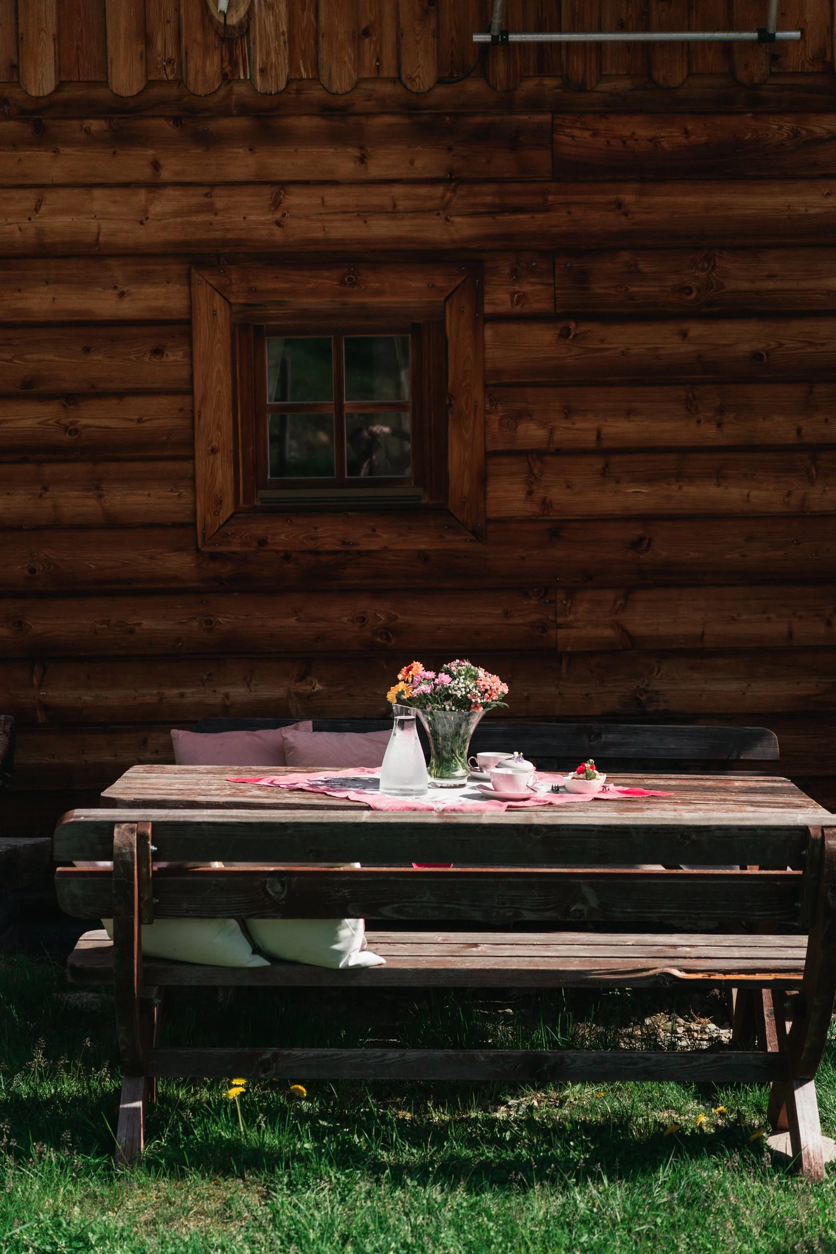 Holzbank mit Tisch darauf ein Blumenstrauß, Kaffee und Dessert vor der Almhütte.
