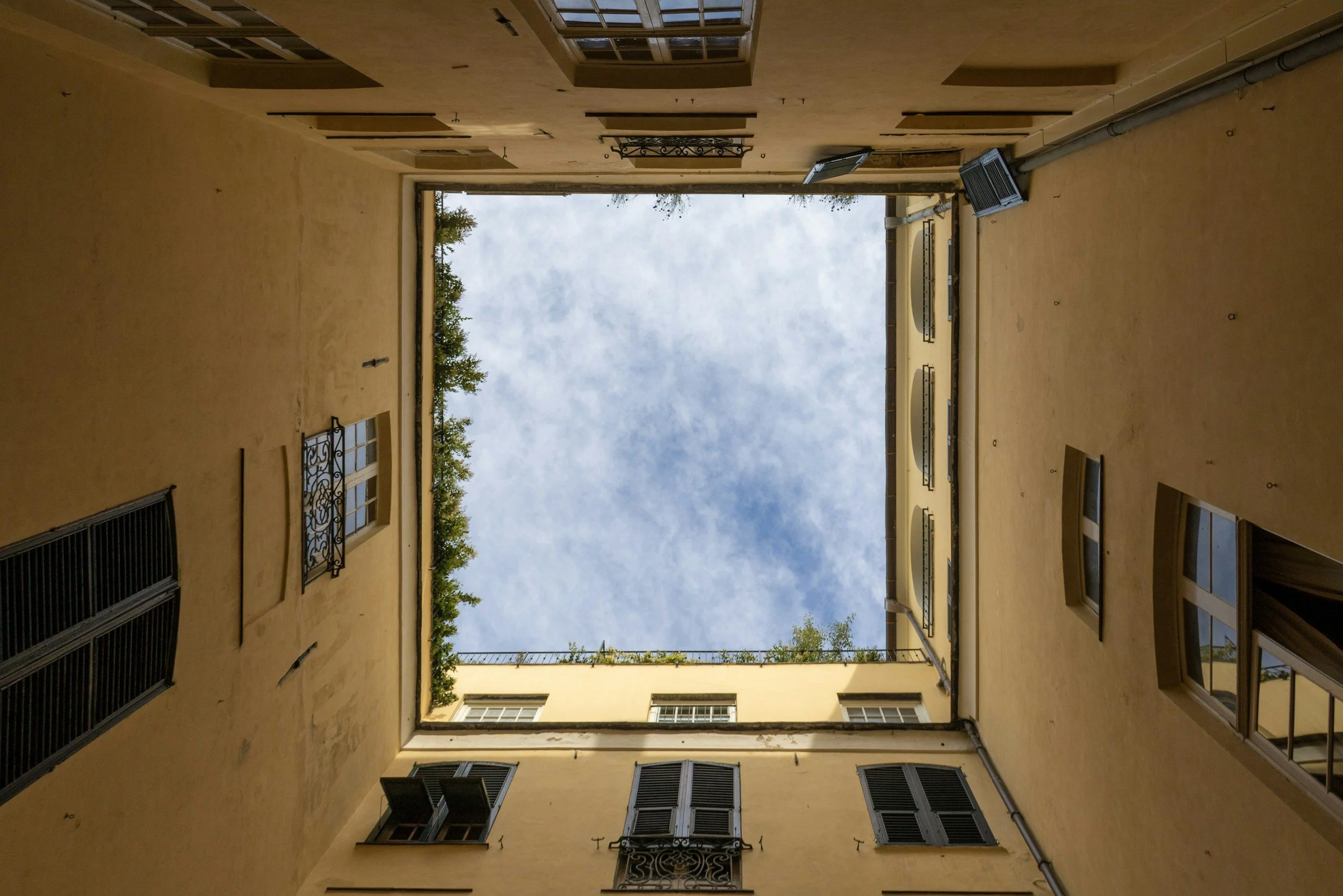 Veduta dal basso verso l'alto di un cortile interno di una palazzina con pareti gialle e molte finestre con persiane, e un cielo nuvoloso visibile dall'apertura superiore.