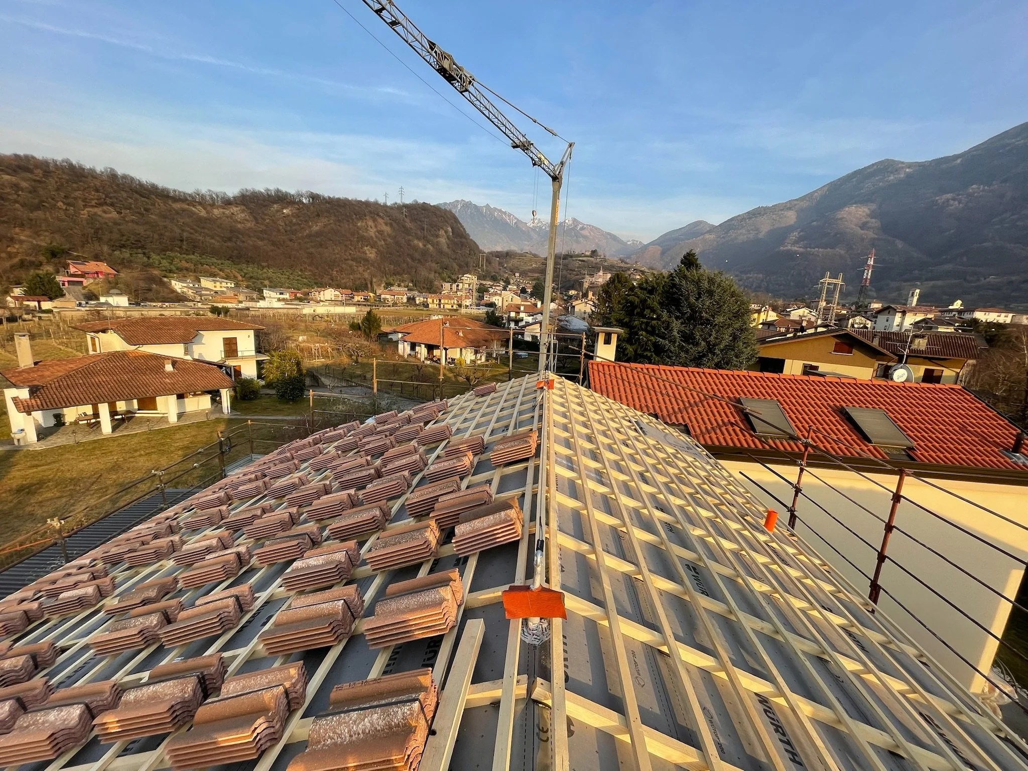 Tetto in costruzione con alcune tegole già posate e altri in attesa, con una gru che lavora sul tetto, in un'area abitata con case e montagne sullo sfondo.