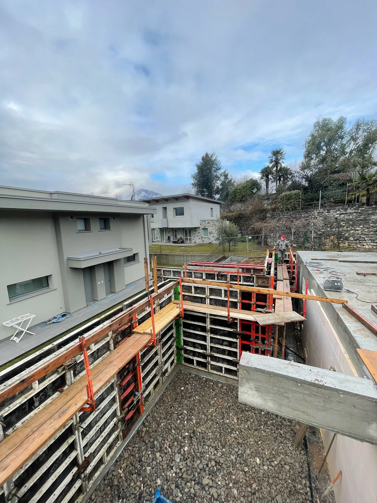 Cantiere di costruzione con strutture di supporto in metallo e legno, operai che lavorano, ed edifici residenziali circostanti con cielo nuvoloso.