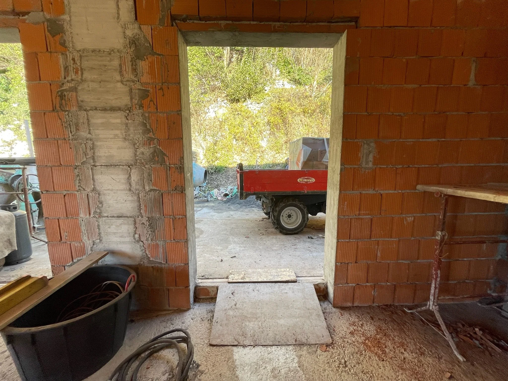 Vista di un'area di cantiere con pareti in mattoni rossi non ancora rifinite, con una porta aperta che dà su uno spazio esterno con un'auto e una piccola motocarrozza rossa.