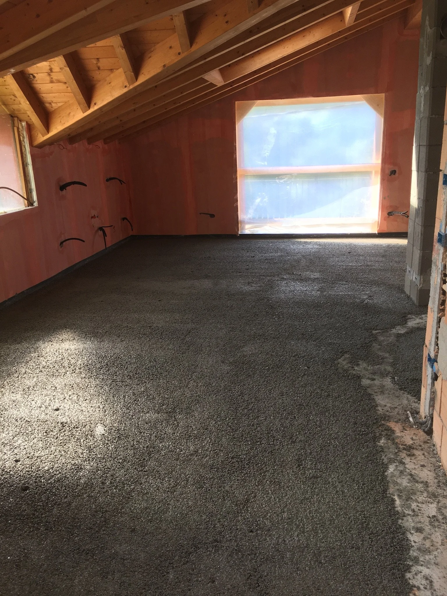 Interno di un attico in costruzione con pavimento in calcestruzzo appena steso, pareti dall'isolamento in fase di finitura e soffitto in legno con travi a vista; finestra grande con vista sull'esterno.