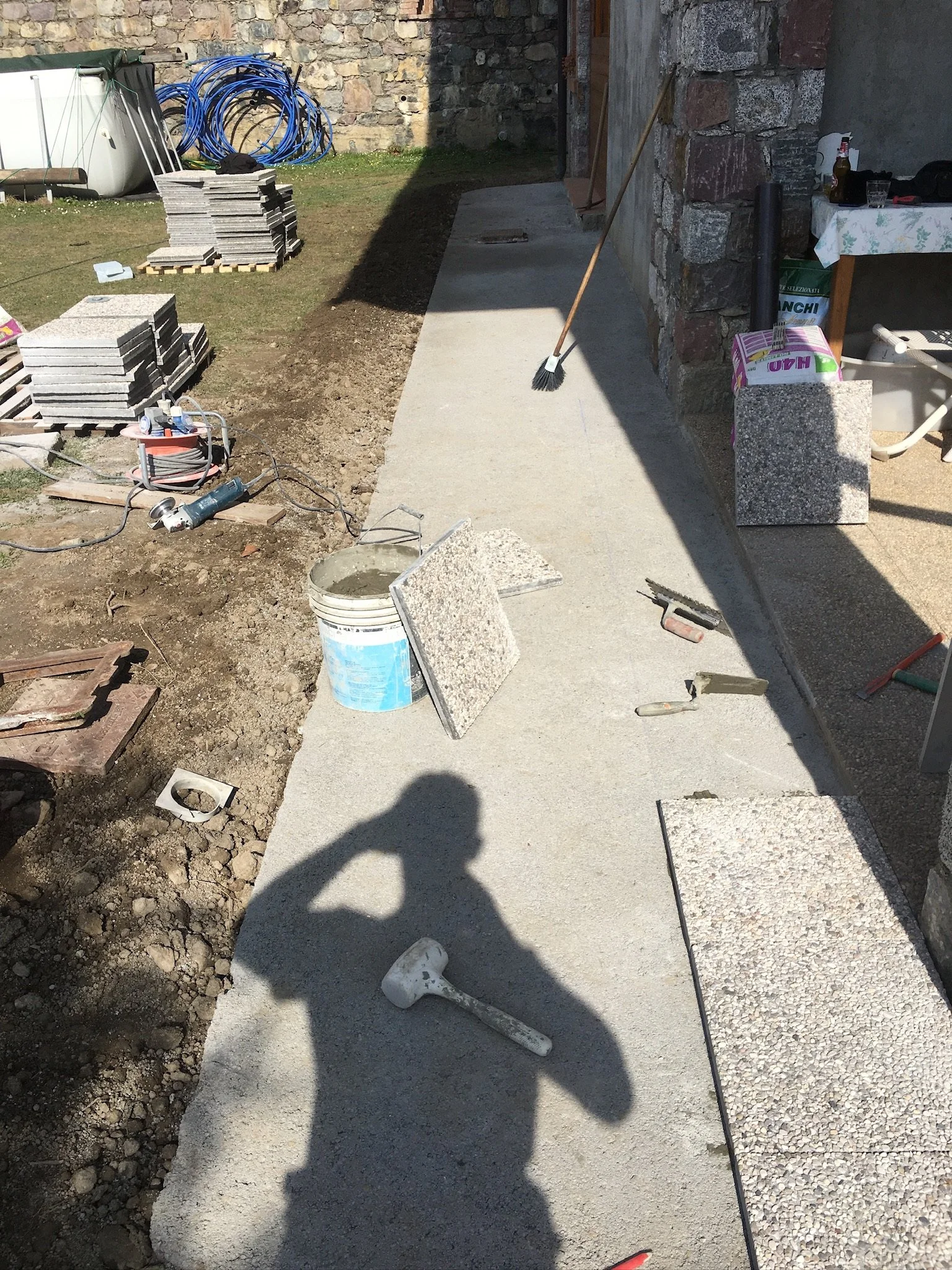 Lavori in corso per la posa di un camminamento in cemento con attrezzi e materiali da costruzione, come secchio, spatole, mattoni e spazzola, in un cortile.