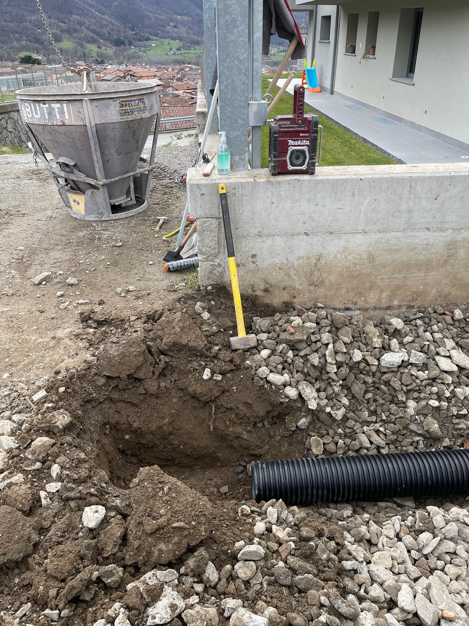 Cantiere edile con buca nel terreno, tubi di plastica nero e attrezzi da lavoro. Sullo sfondo vedono un edificio, un balcone con alcuni oggetti e la vista di un paesaggio collinare.