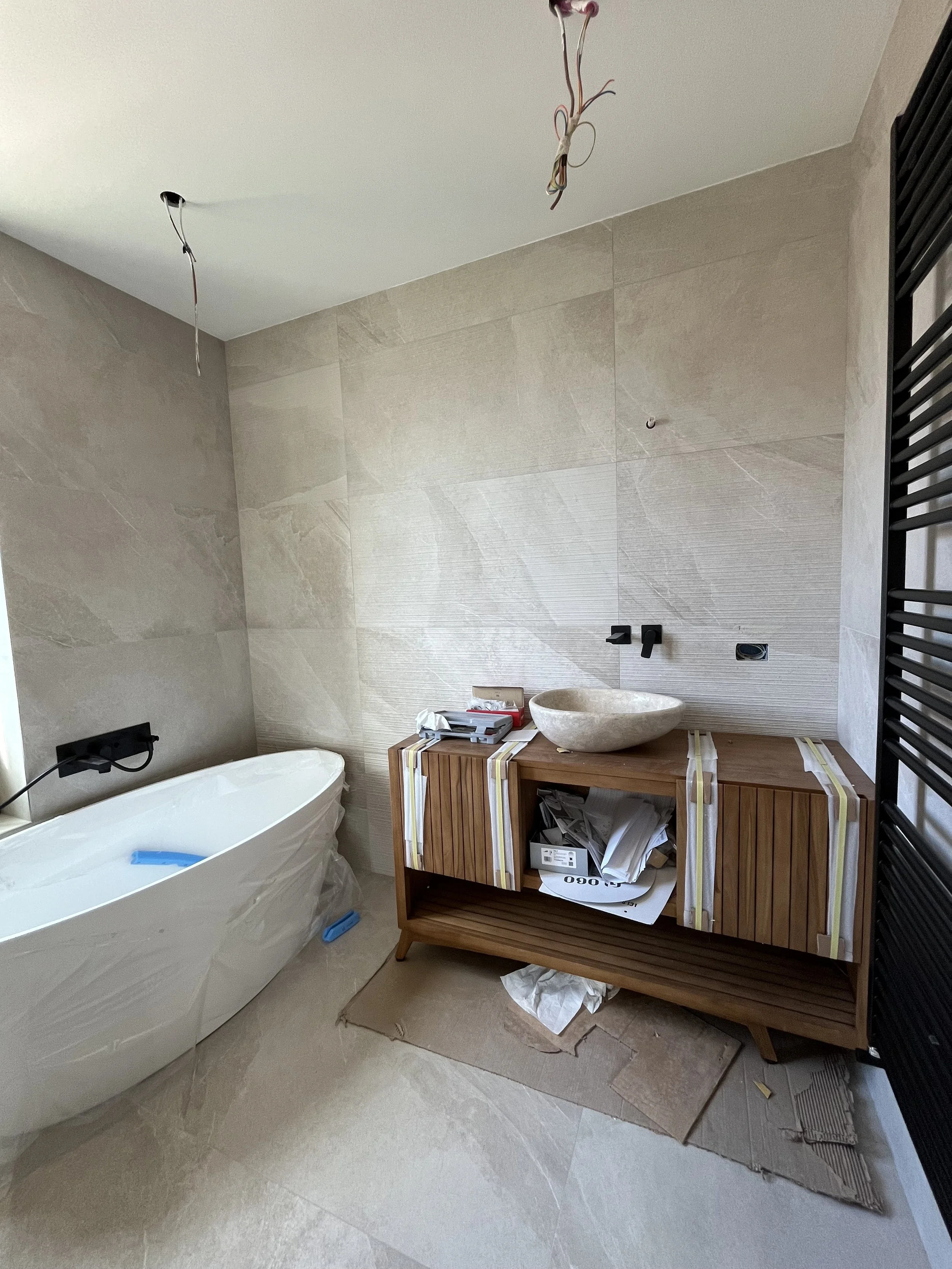 Stanza di bagno in fase di ristrutturazione con vasca da bagno, mobile con lavabo in pietra, pareti e pavimenti in piastrelle beige, fili elettrici scoperti e scatole di attrezzi e materiali.