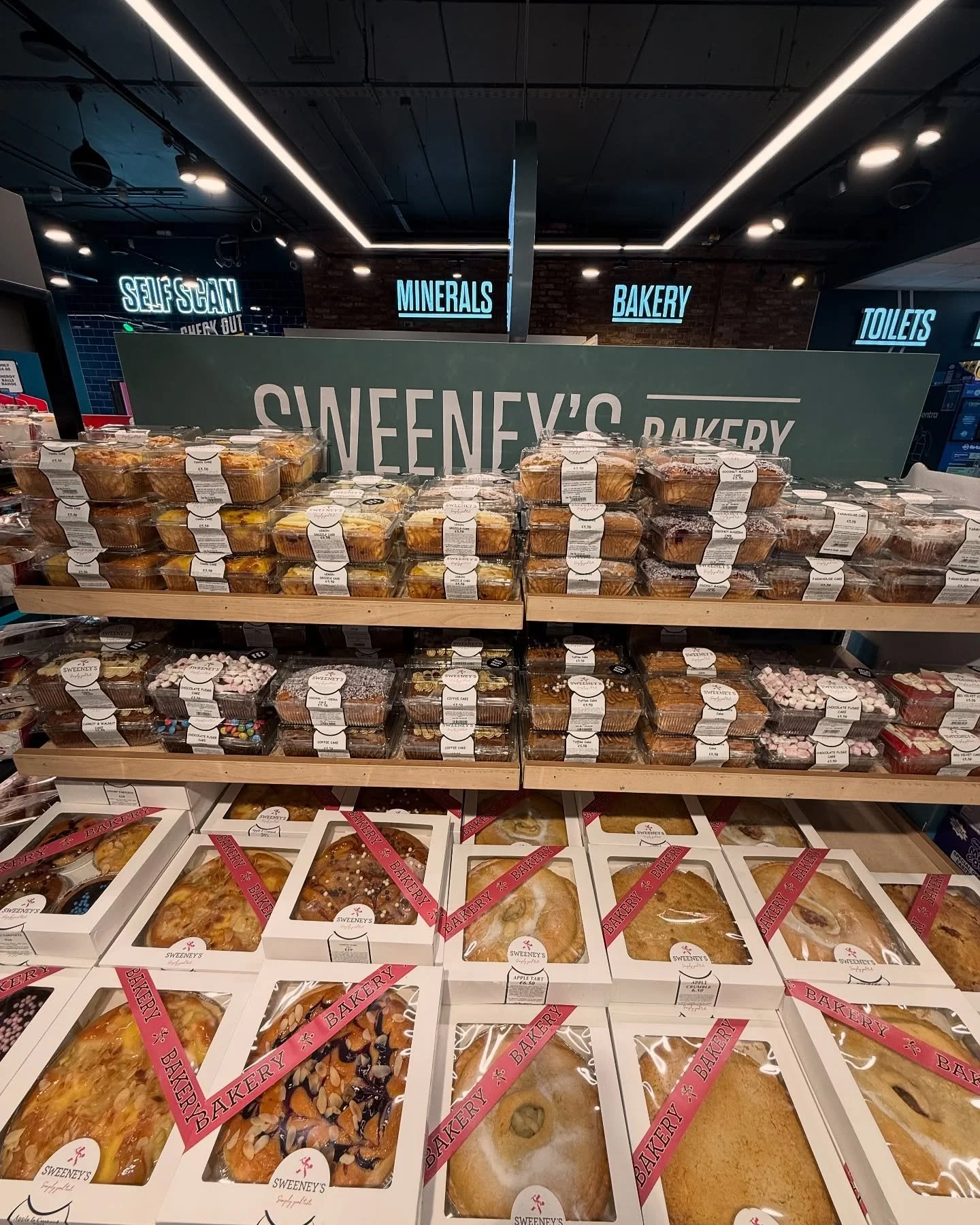 Display of bakery items in packaged boxes at Sweeney's Bakery in a supermarket, with signs for 'Self Scan,' 'Minerals,' 'Bakery,' and 'Toilets' in the background.