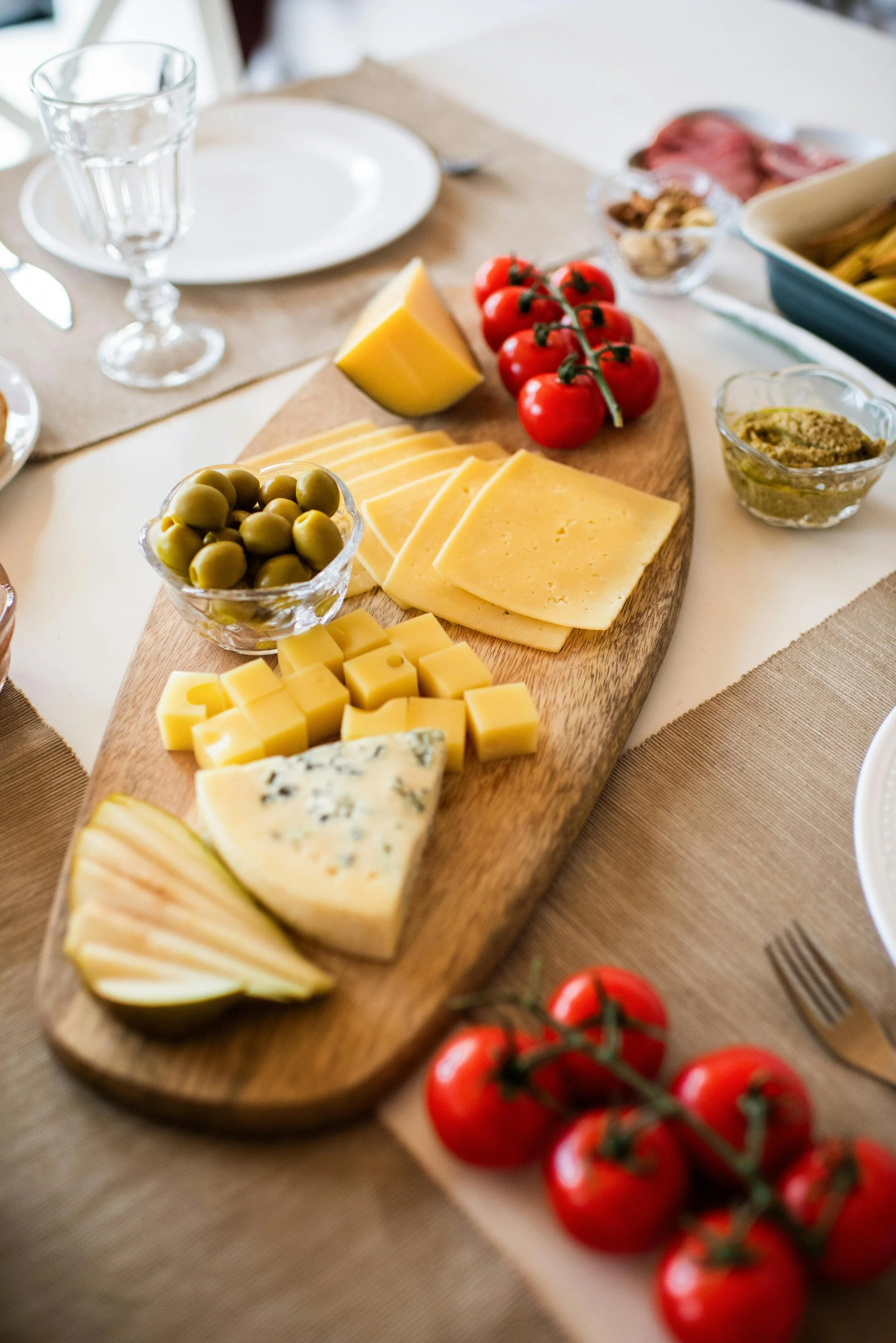 A wooden serving board with an assortment of cheeses, green olives, cherry tomatoes on the vine, and a small bowl of green spread. The background includes a table setting with a glass, a plate, and other dishes.