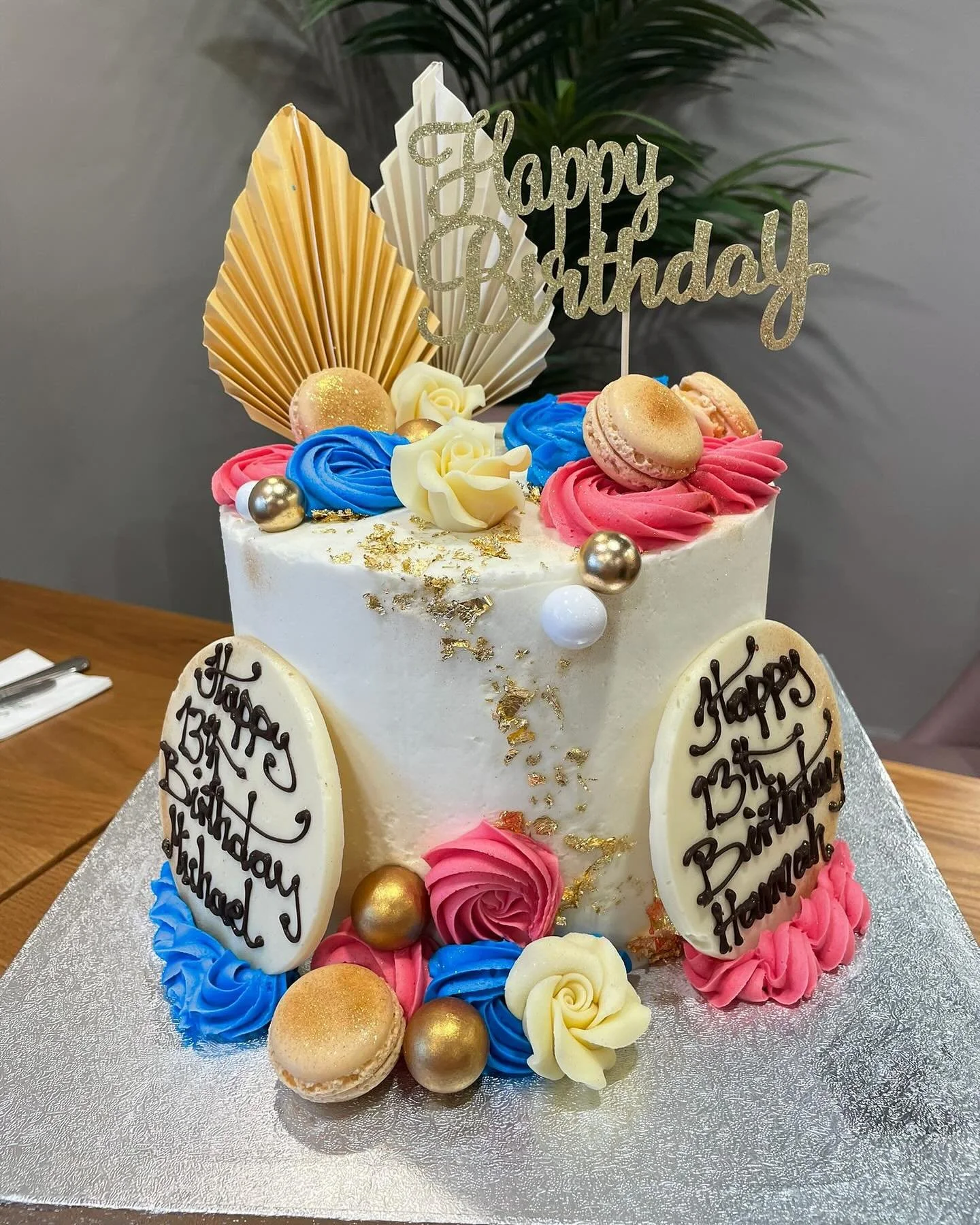 A birthday cake decorated with pink, blue, and white frosting flowers, macarons, gold and white decorative balls, gold leaf accents, and a glittery 'Happy Birthday' topper. The cake is also adorned with two white chocolate plaques with black script writing.