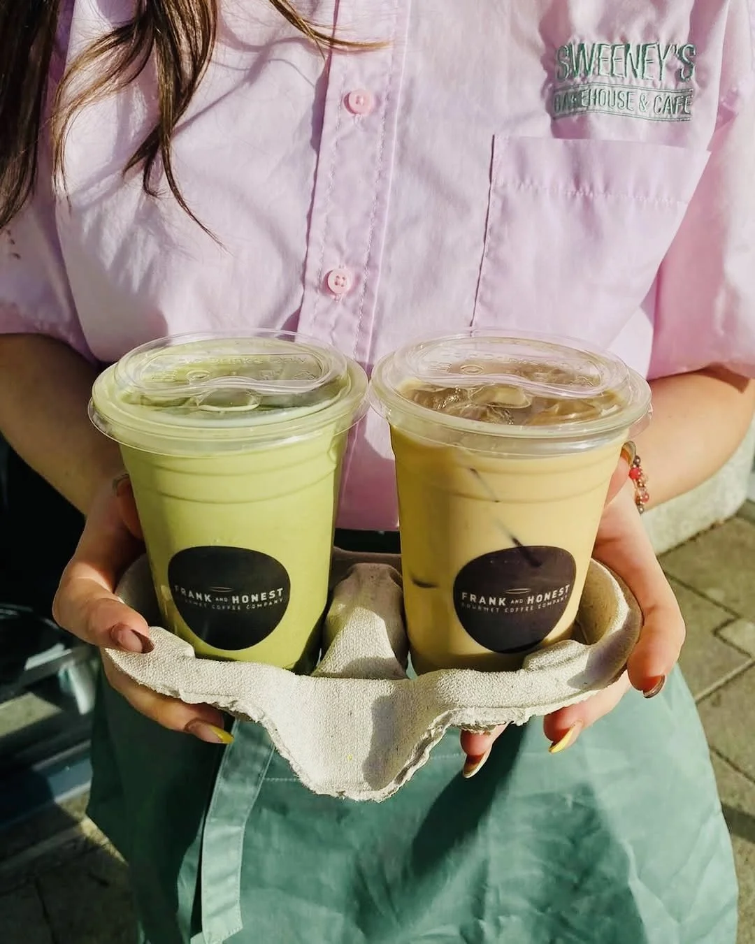 A person in a pink shirt holding a tray with two iced coffee drinks in clear cups, one green and one cream-colored, from Frank and Honest. The cups have black logos and are covered with plastic lids.
