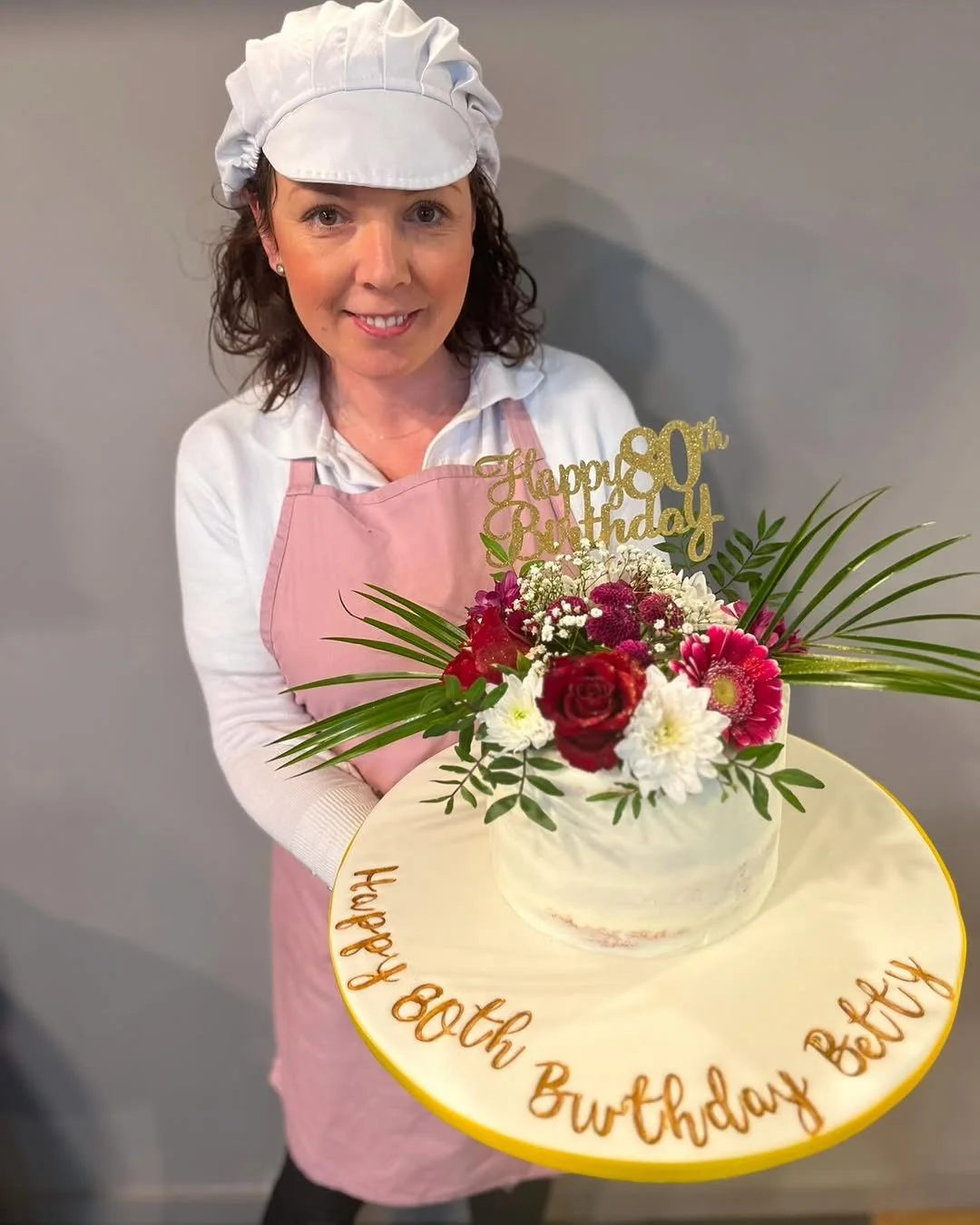 A woman dressed as a chef holding a birthday cake with flowers and a gold 'Happy 80th Birthday Granny' topper.