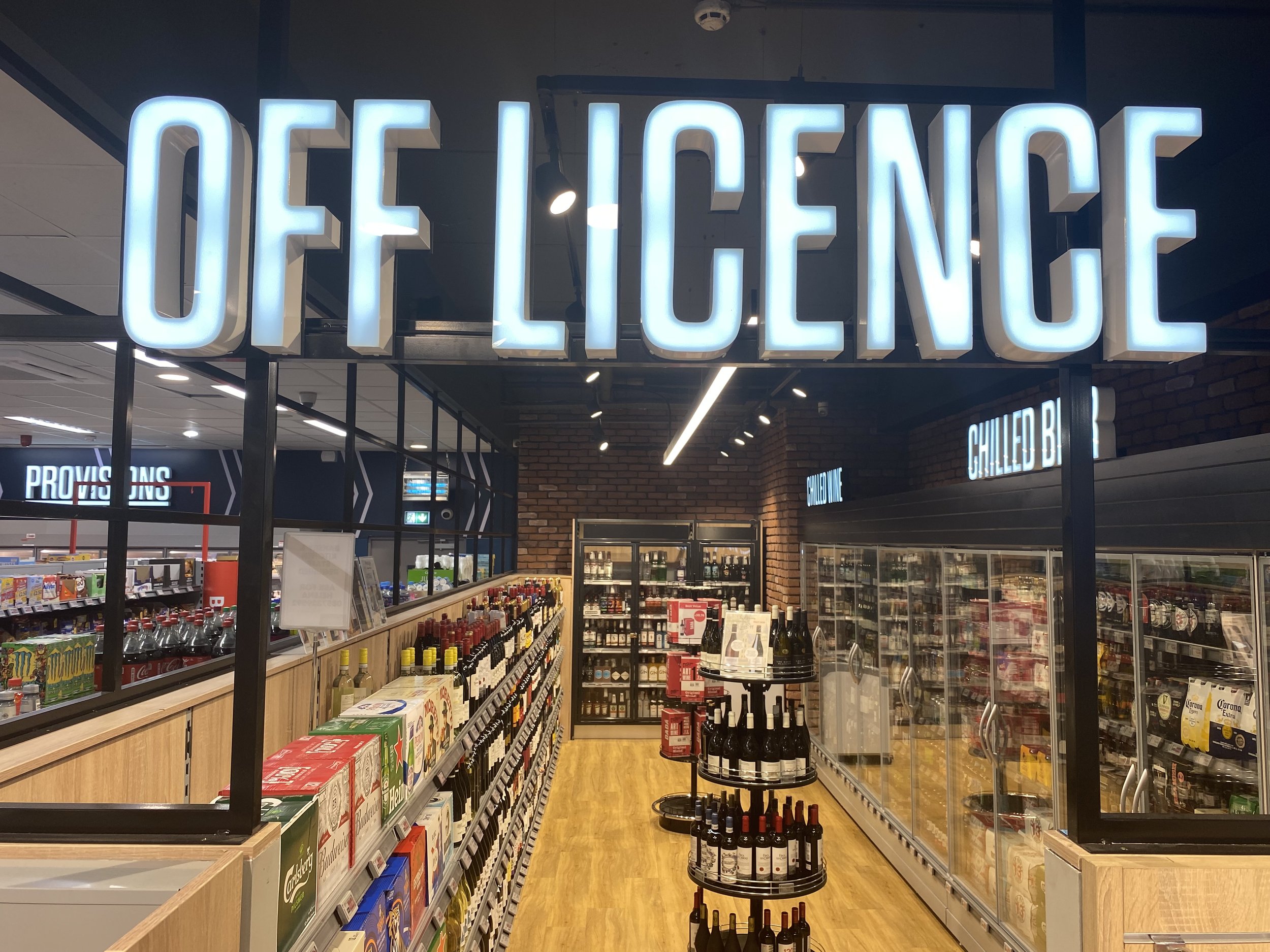 Inside a store with a large illuminated sign that reads 'OFF LICENCE, shelves stocked with wine bottles, refrigerated beverage cases, and snack items visible.