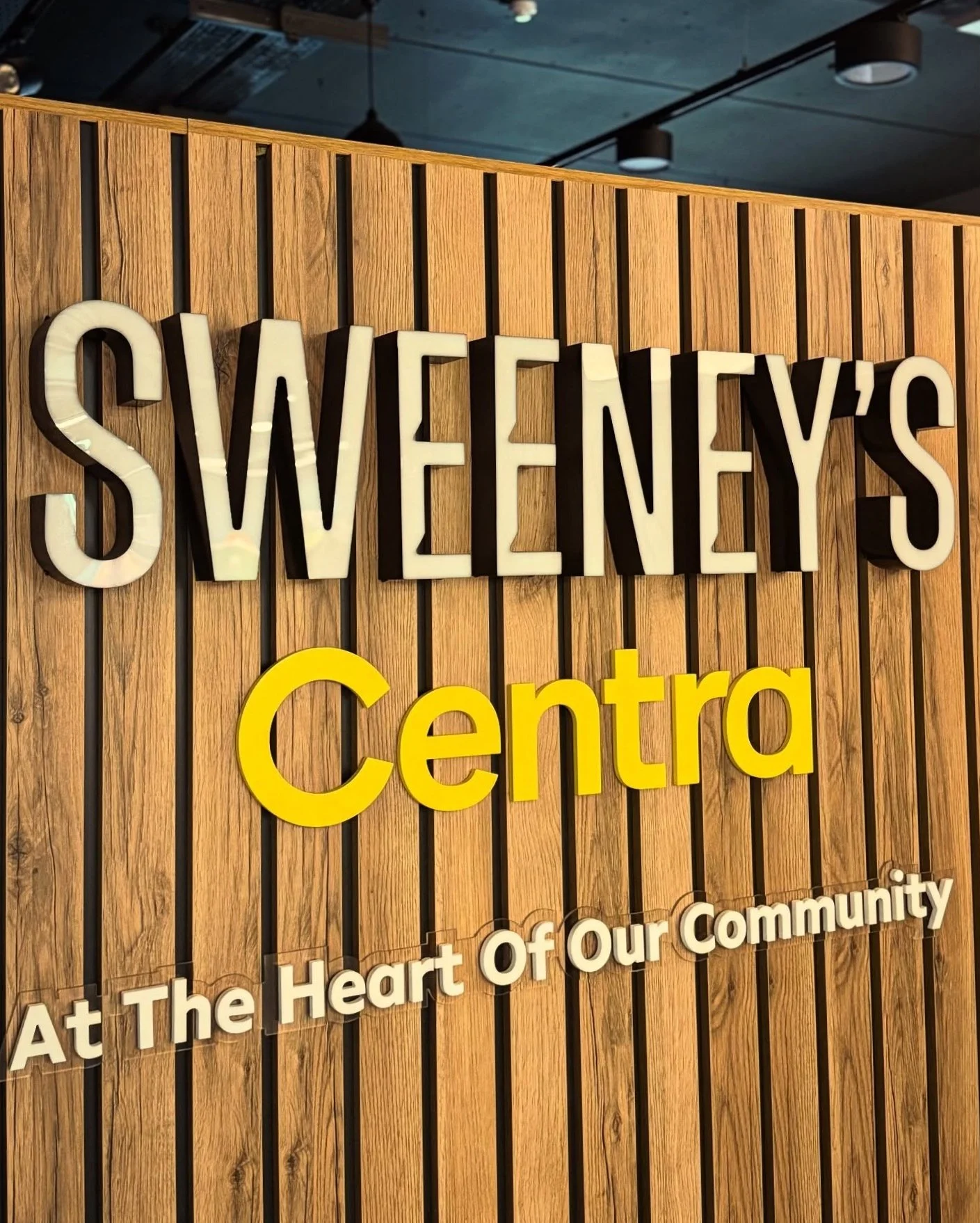 Wooden wall with large white and yellow 3D text reading 'SWEENEY'S Centra' and smaller text below that says 'At The Heart Of Our Community'.