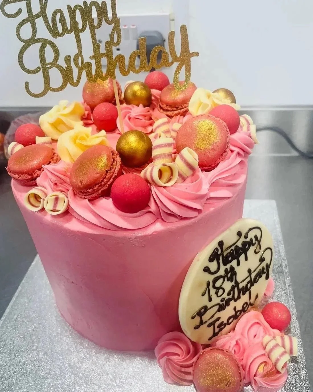 Pink birthday cake decorated with macarons, chocolate balls, and white and pink swirl decorations, with a "Happy Birthday" topper and an oval white chocolate plaque reading "Happy 18th Birthday Izdee."