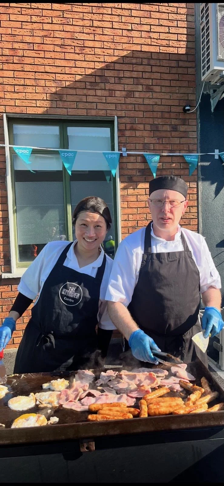 Two chefs, a woman and a man, cooking breakfast on a large outdoor griddle. The woman is smiling and the man appears focused. They are wearing aprons and gloves. There are eggs, bacon, and sausages on the griddle, with a brick building and window in the background.