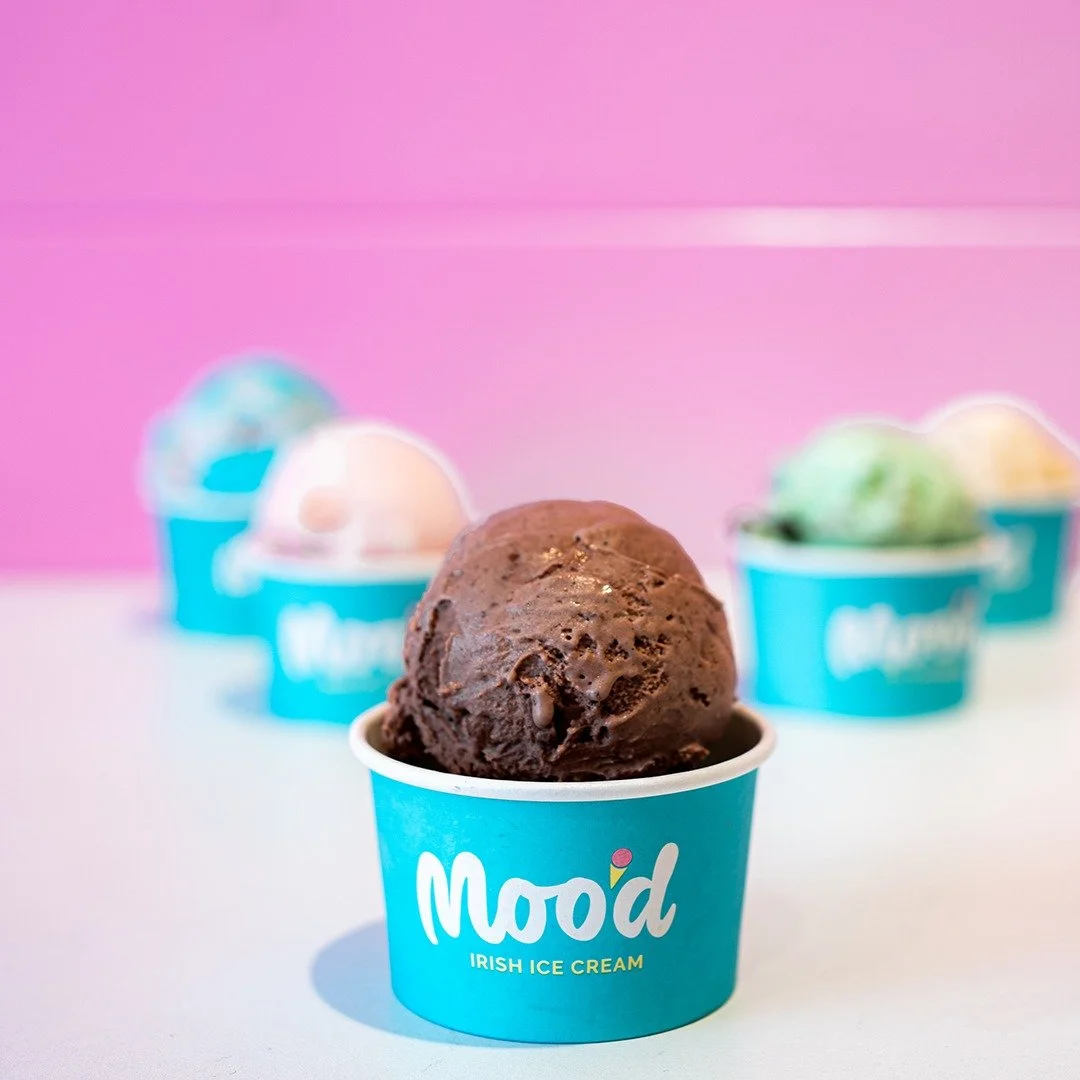 A scoop of chocolate ice cream in a blue cup with the label 'Mood Irish Ice Cream,' with three other colorful ice cream cups blurred in the background against a pink wall.