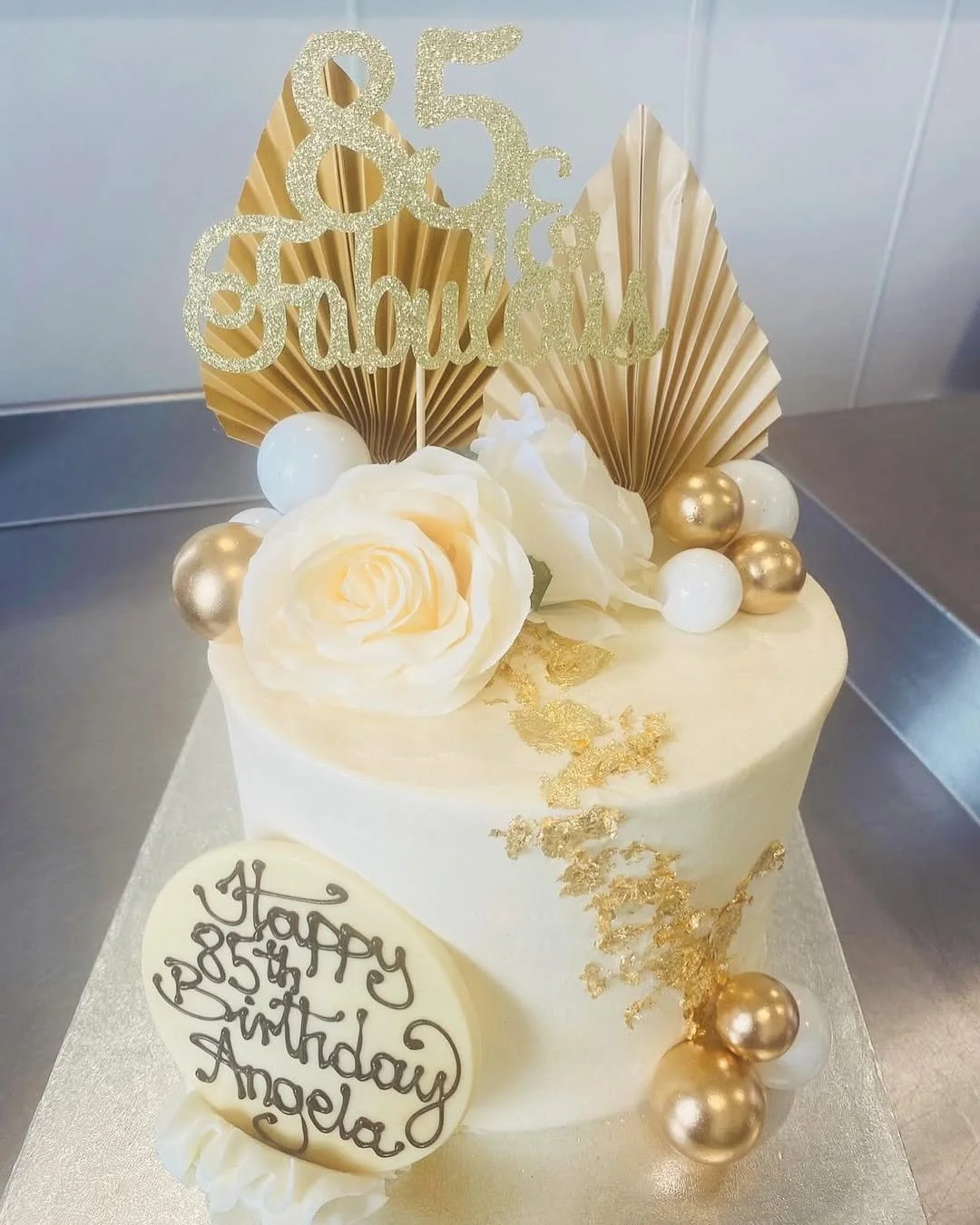 A white birthday cake decorated with gold and white balloons, white roses, gold leaf accents, and a glittery gold topper that reads '85 & fabulous.' A white oval plaque with black cursive writing says 'Happy 85th Birthday Angela.'