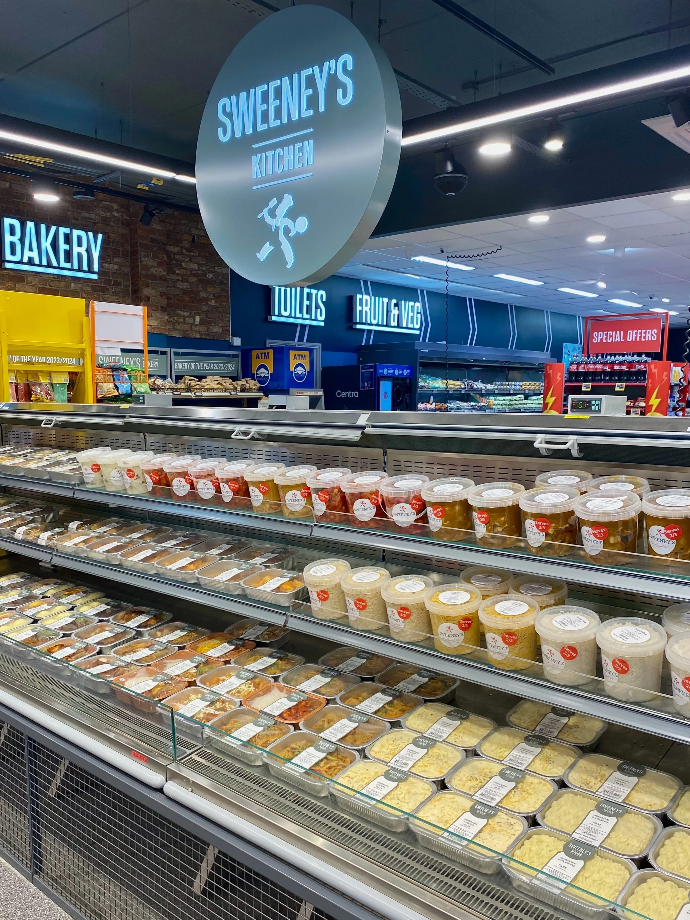 A grocery store refrigerated section with prepackaged cheeses and dips. Signage above indicates sections for bakery, toilets, fruit and vegetables, and Sweeny's Kitchen, with illuminated blue signs in the background.