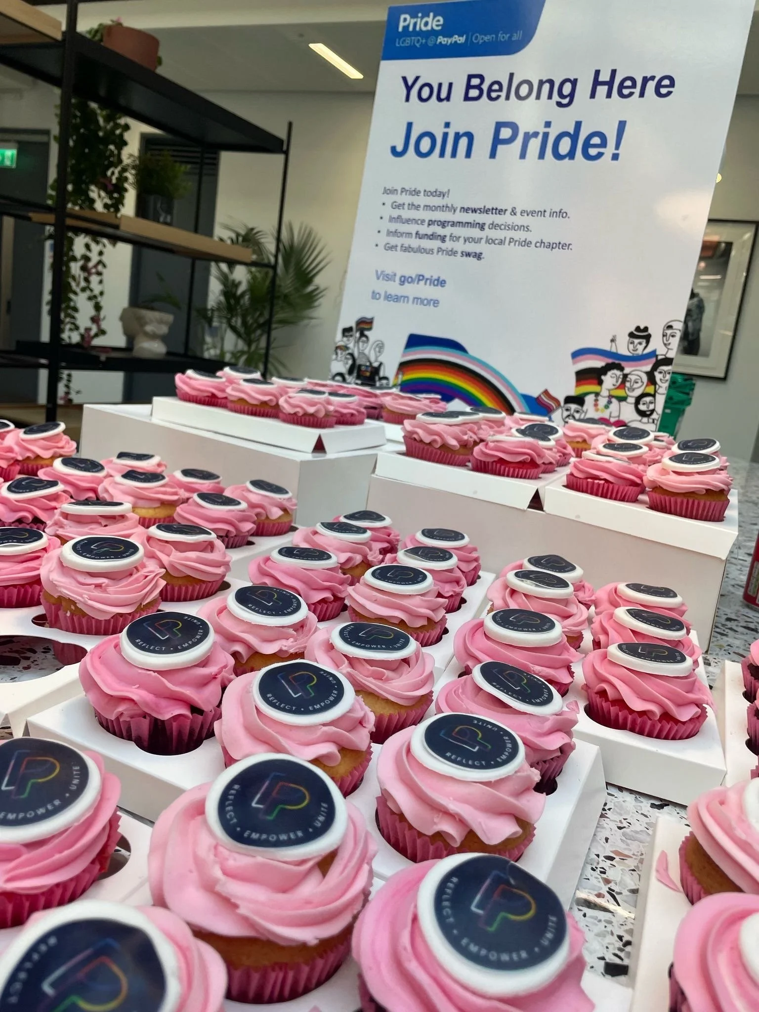 Pink cupcakes adorned with round stickers featuring a colorful 'L' logo and the words 'Reflect, Empower, Unify.' A Pride sign in the background inviting people to join Pride, with a rainbow graphic and illustrations of diverse people, promotes LGBT+ community support.