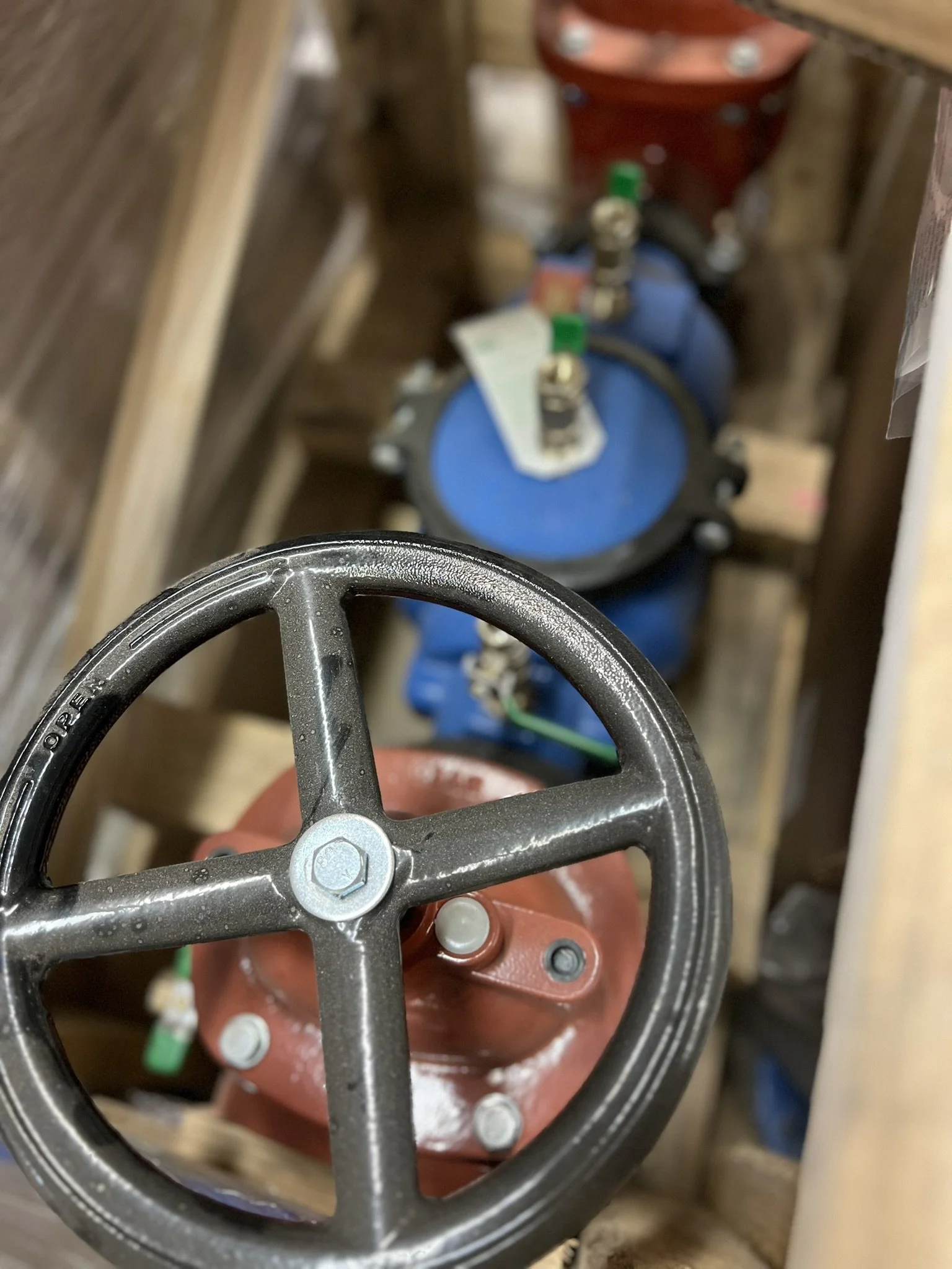 Close-up of a black valve wheel with a wooden handle, part of a blue tank with a red valve and green and black fittings, in a wooden storage area.