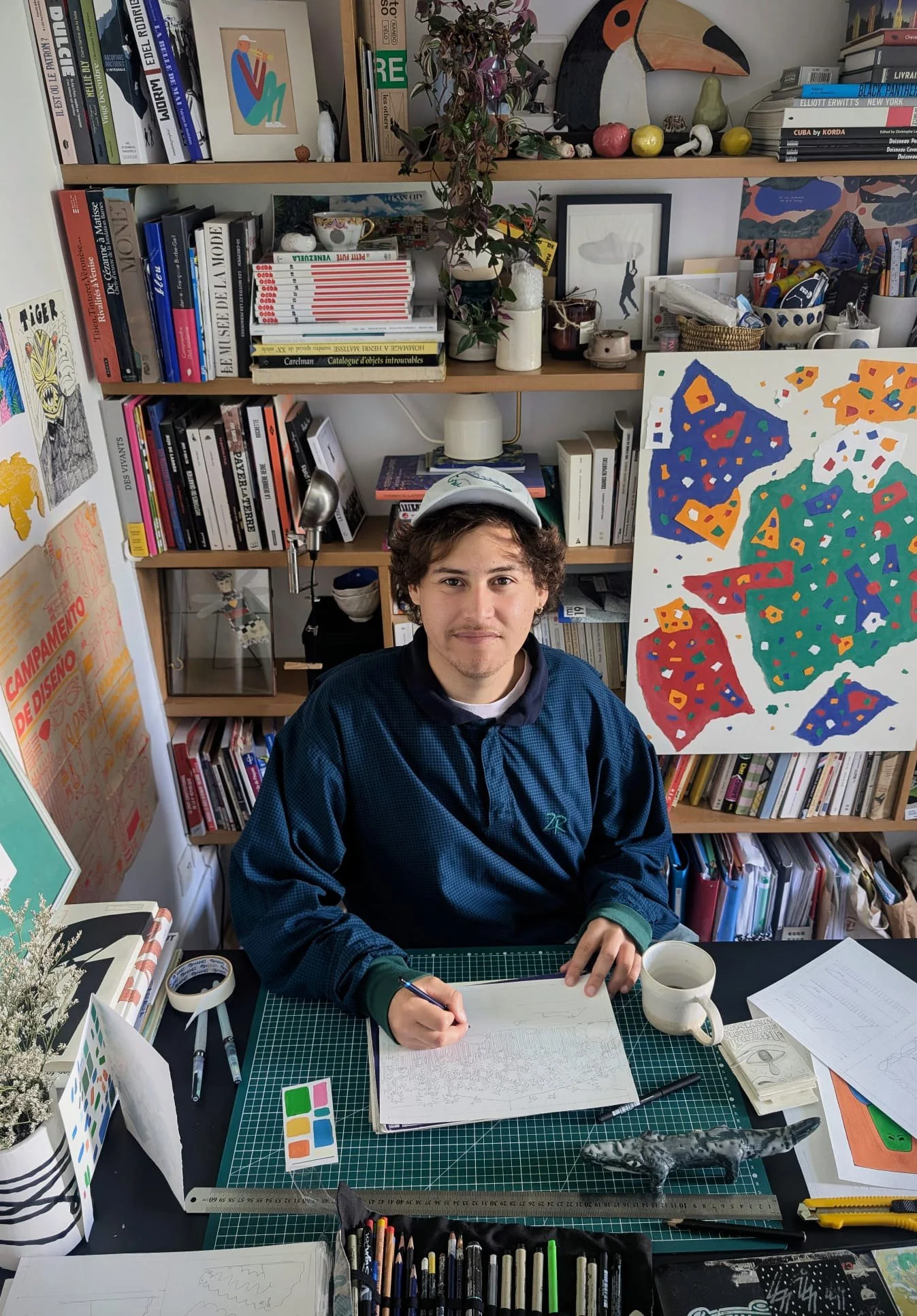 A young man with curly brown hair, wearing a cap and a blue jacket, is sitting at a cluttered desk in a room filled with books, art supplies, and colorful artwork. He is holding a pen and working on a large sketchbook, surrounded by various art tools and a white mug.