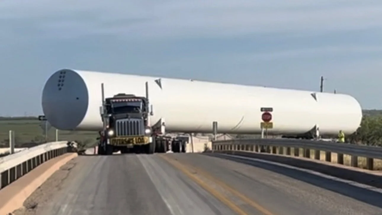 Truck transporting large cylindrical load on highway, labeled "oversize load," with clear sky in background.