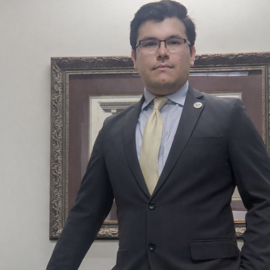 Man in a dark suit with glasses, light blue shirt, and yellow tie standing indoors in front of a framed picture.