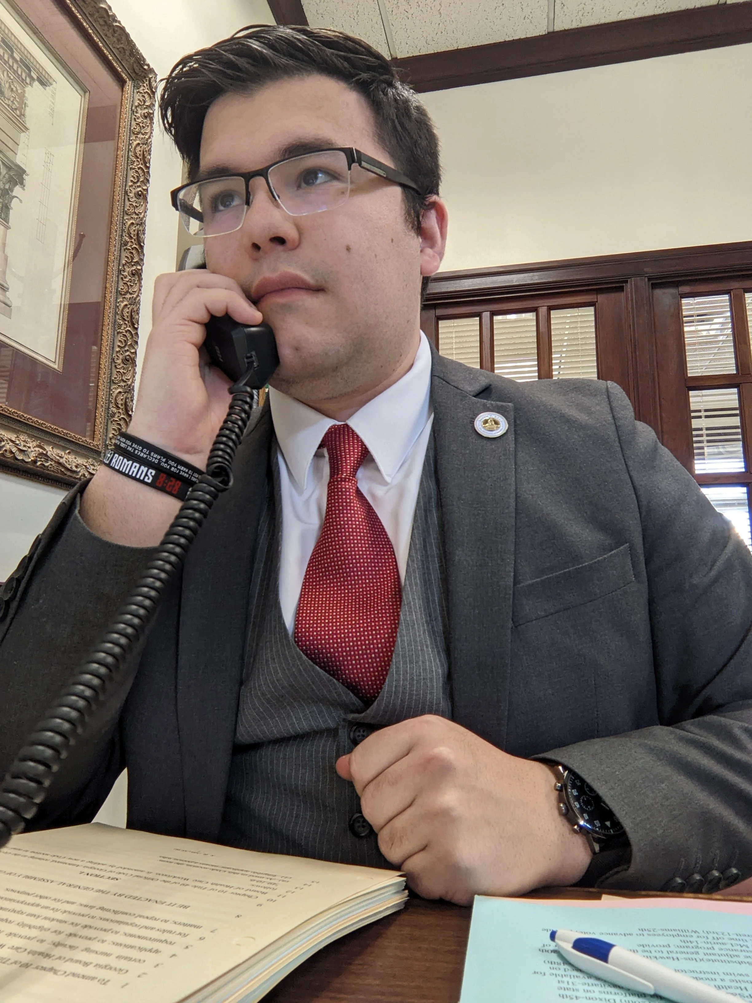 Christopher Carrusca wearing glasses, a red tie, and a wristwatch, is sitting at a desk talking on a landline phone. There are documents and a pen on the desk, and a framed picture on the wall behind him.