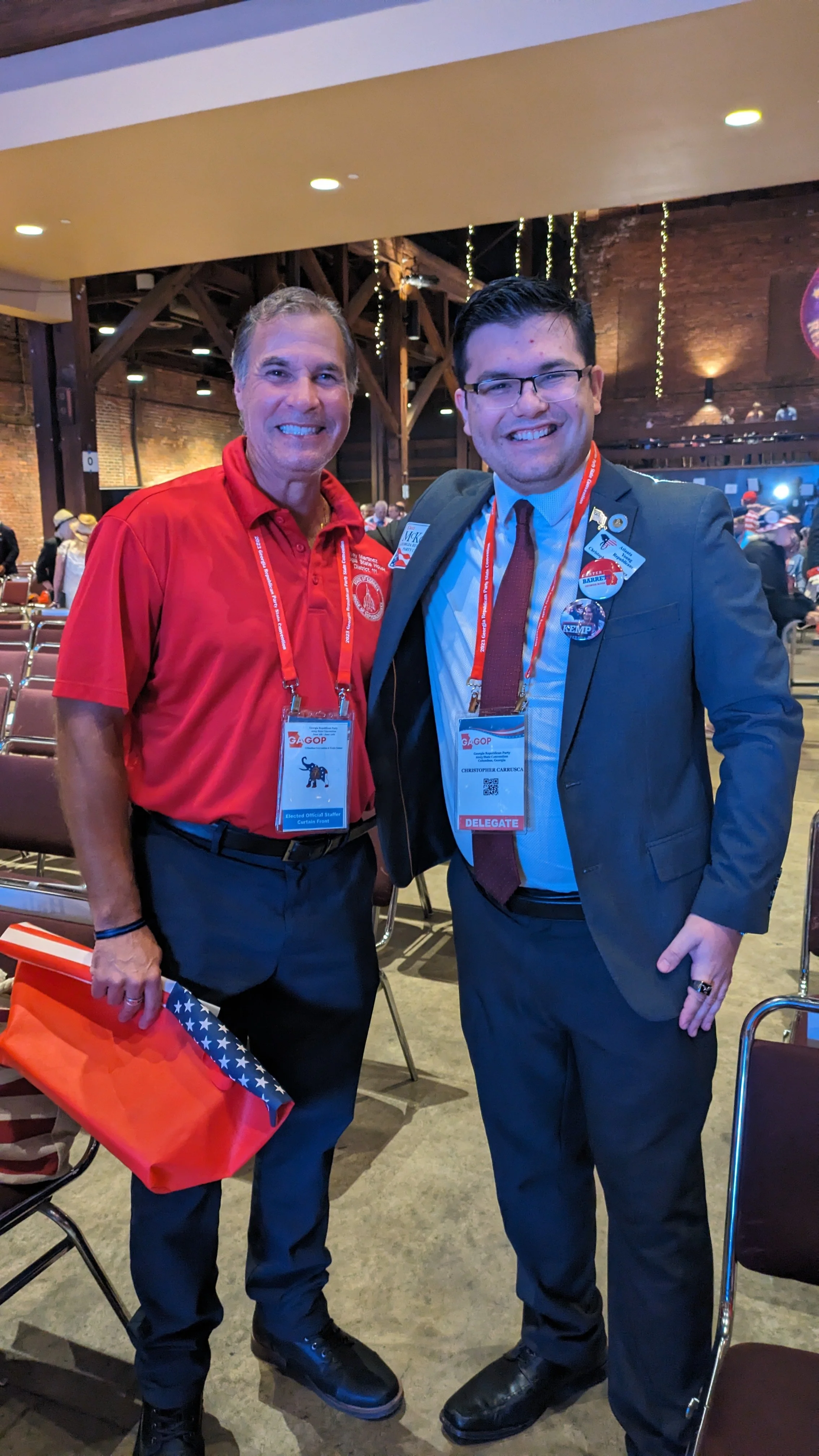 Two men standing inside a decorated venue, smiling for the camera. The man on the left is wearing a red polo shirt with a badge that says 'Elected Official Staff' and holding an American flag. The man on the right is dressed in a suit with a name tag