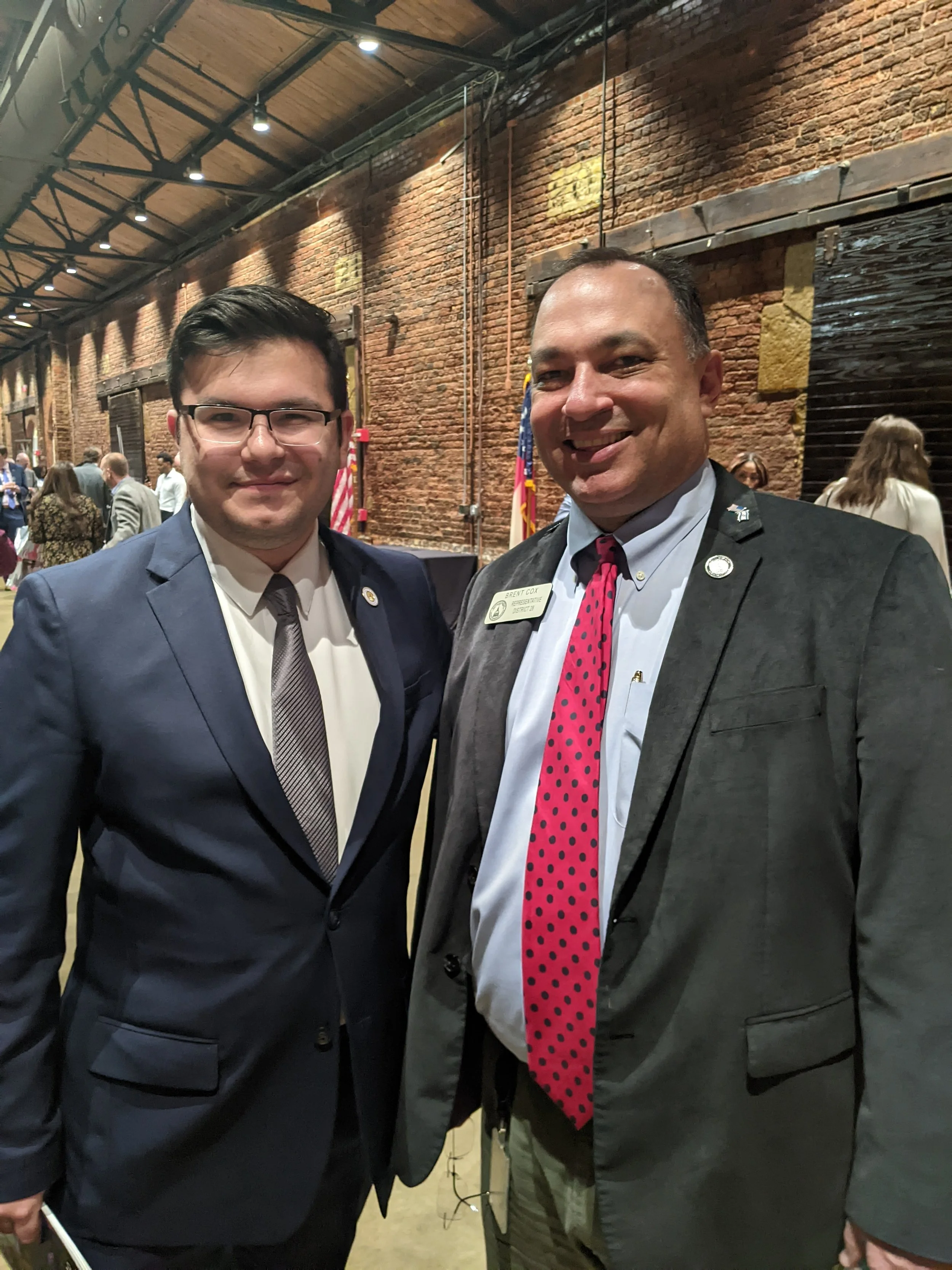 Two men in suits smiling and standing close together in a large, brick-walled event space with other people in the background.