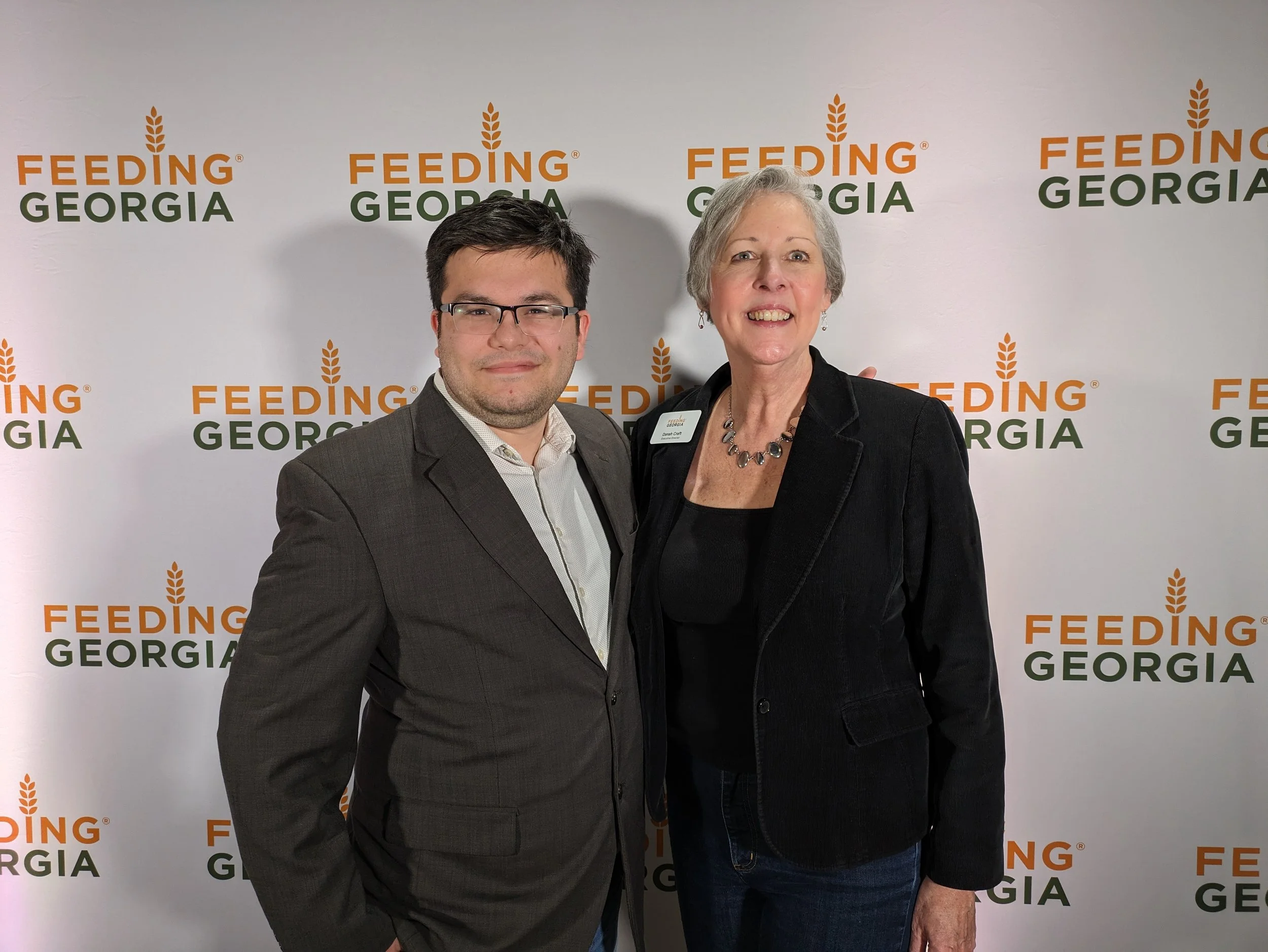 Two people standing in front of a backdrop with the words 'Feeding Georgia' and a wheat design. One is a young man in glasses wearing a suit, and the other is an older woman with short gray hair wearing a black jacket, smiling.