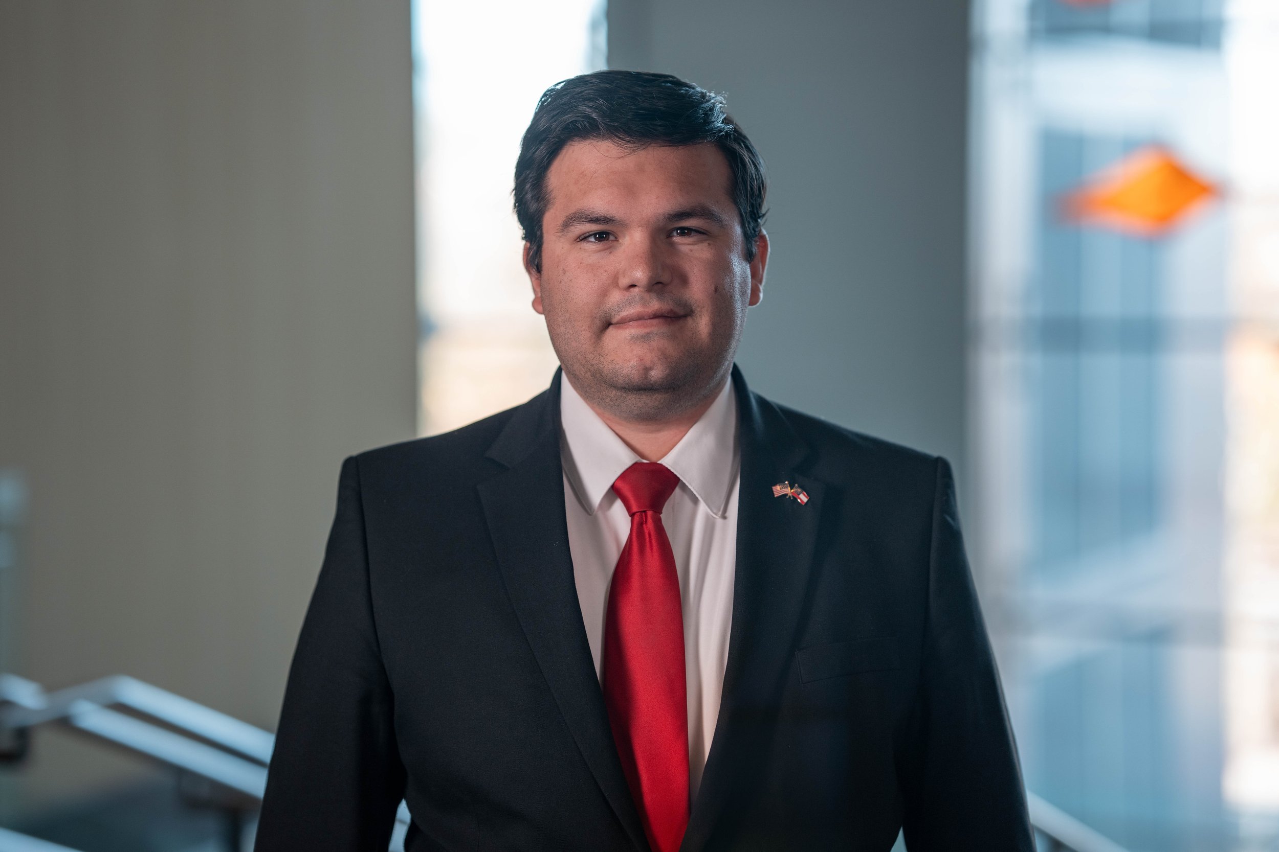 A man in a suit and red tie standing indoors near a window with a blurred background.