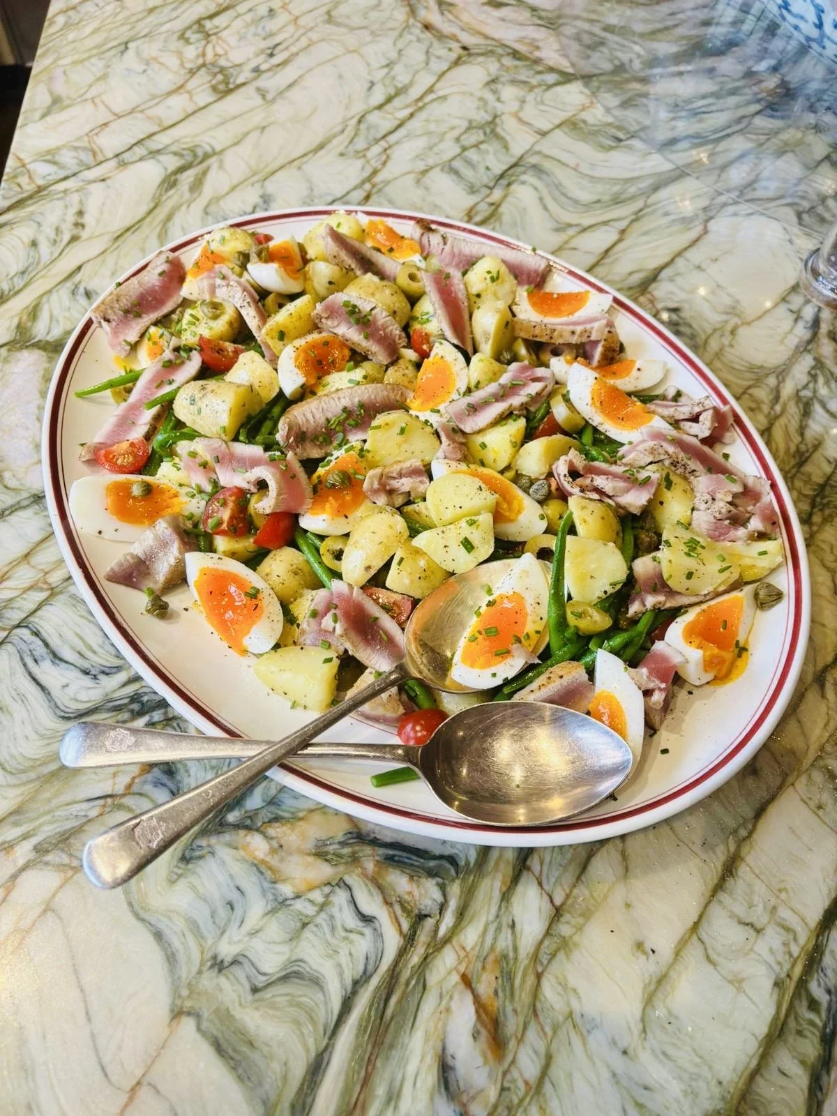 Plate of nicoise salad with seared tuna, potatoes, boiled eggs, green beans, cherry tomatoes, capers, and herbs on a marble table.