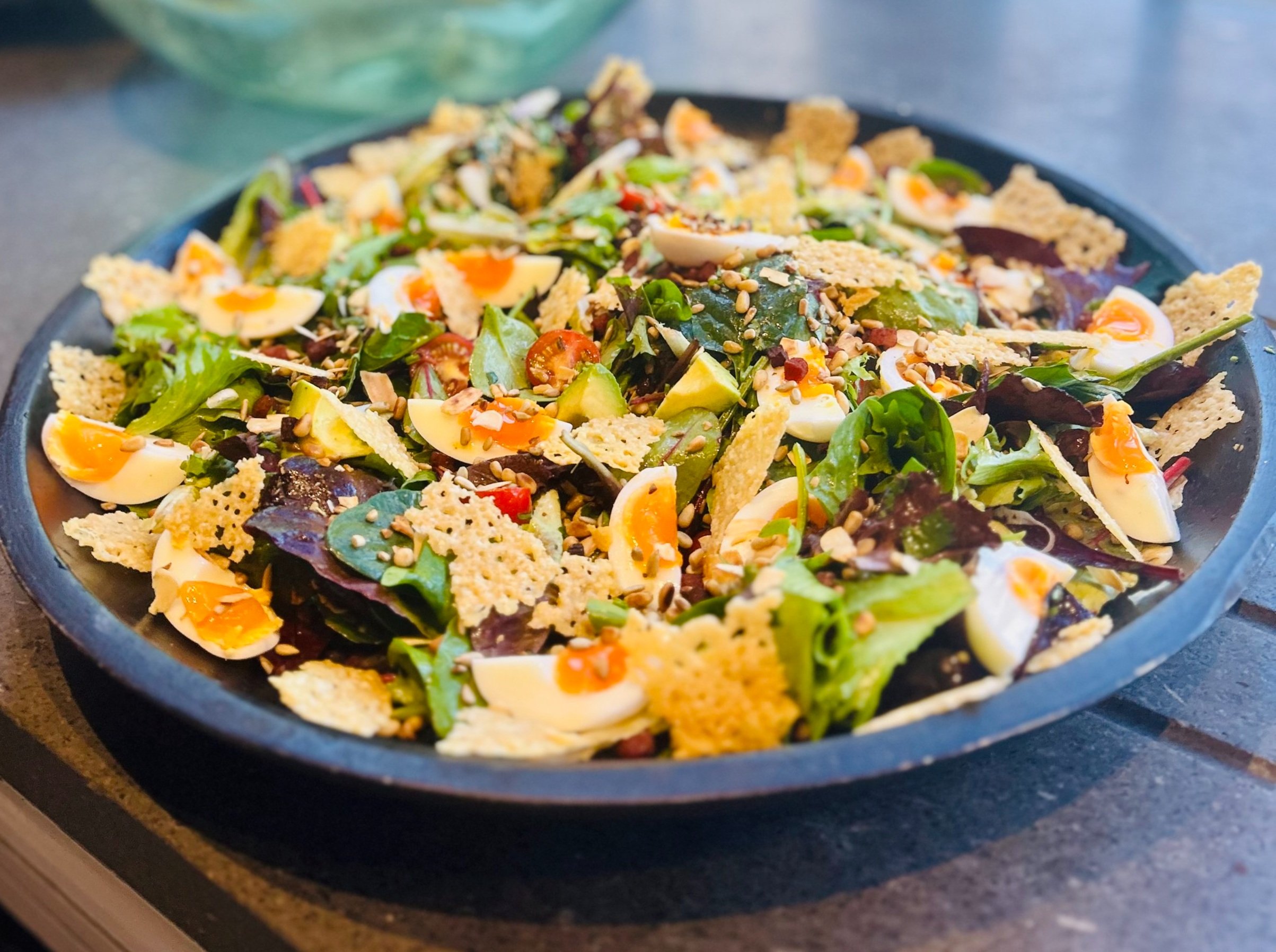Mixed salad with leafy greens, sliced eggs, cherry tomatoes, seeds, and crispy cheese crisps on a dark plate.