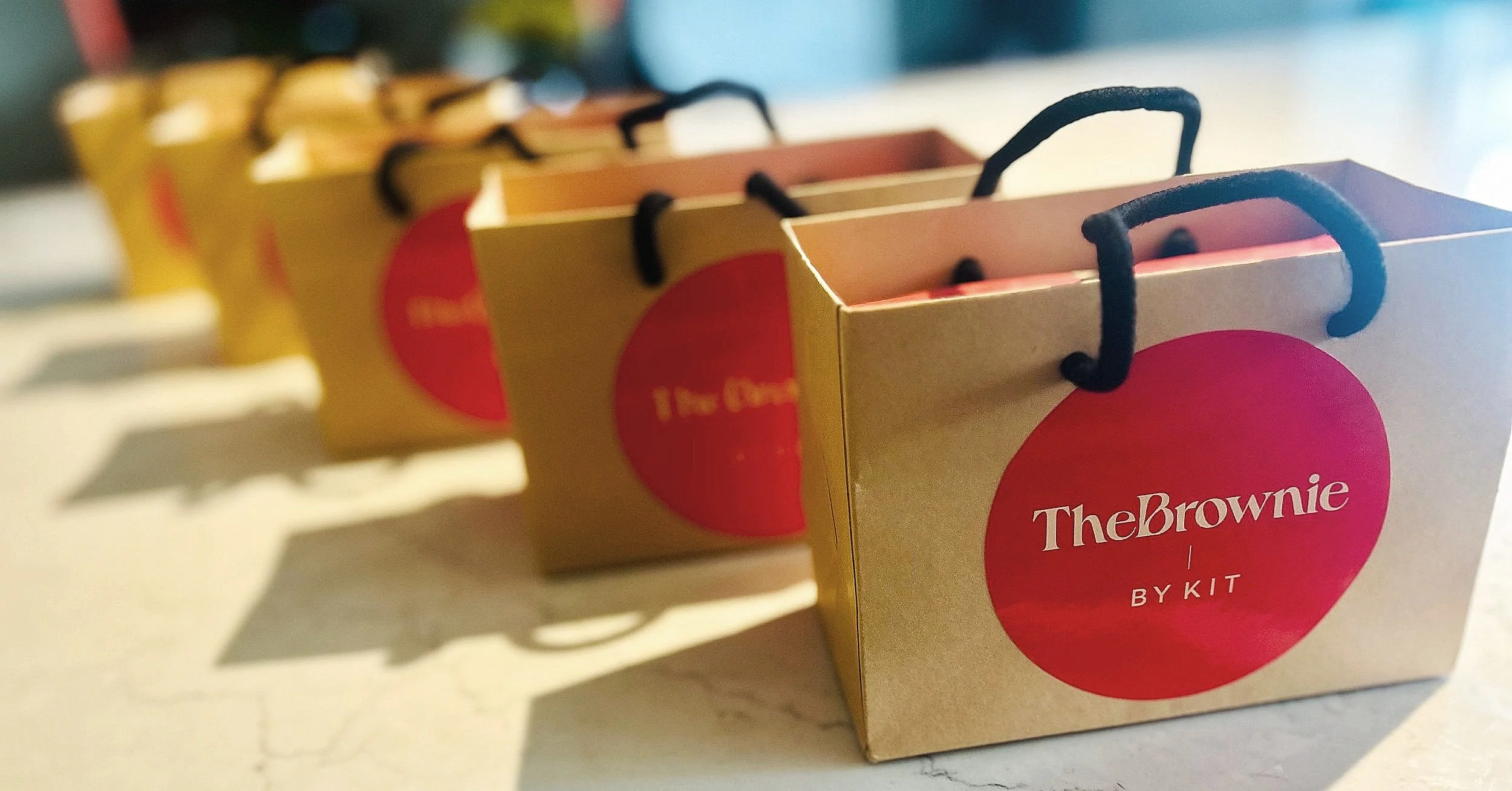 Row of brown paper bags with red circles and text "The Brownie by Kit" on a marble surface