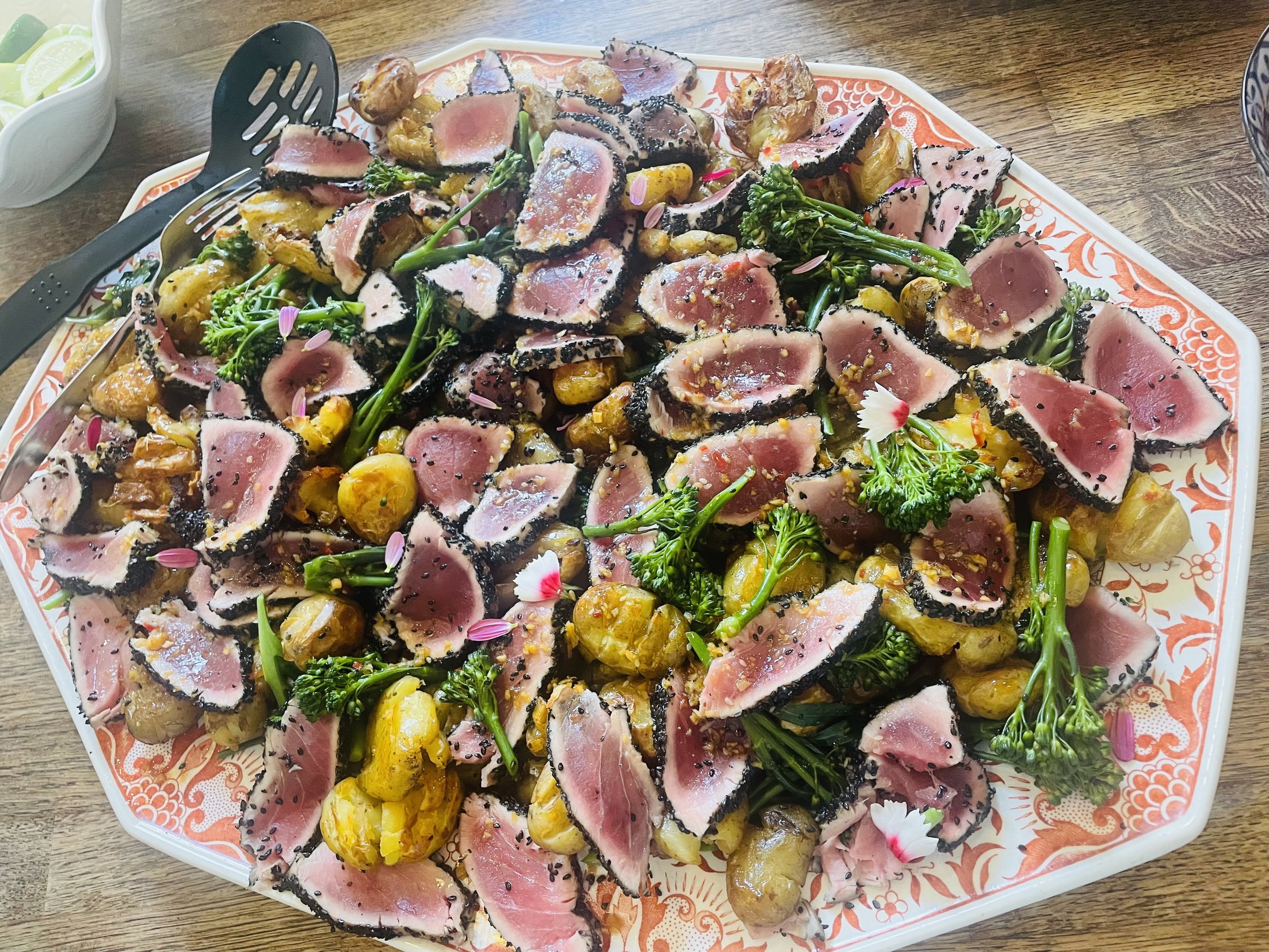 Plate of seared ahi tuna slices with sesame crust, served with roasted potatoes, broccolini, and garnished with flower petals.