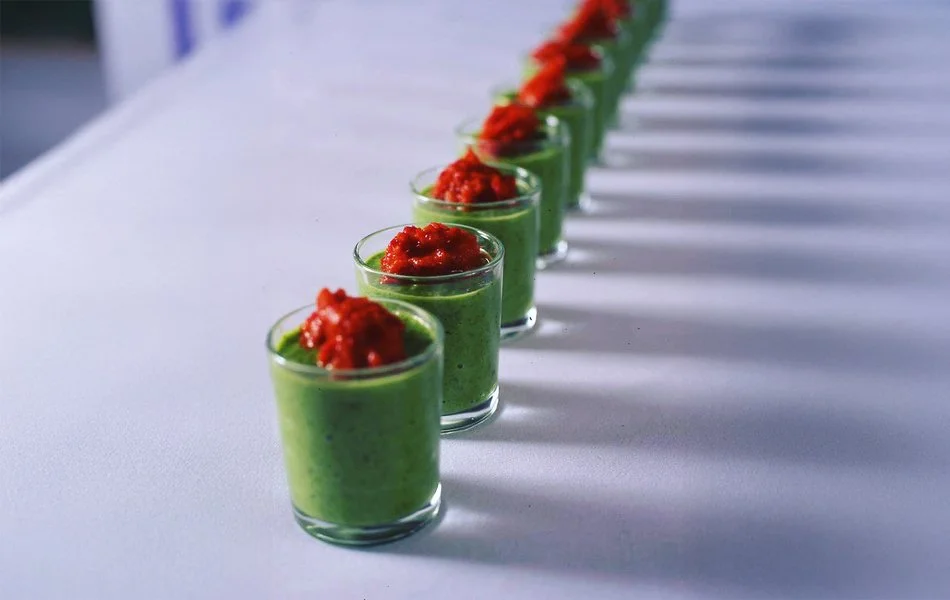 Row of small clear glasses filled with green smoothie and topped with red sauce, on a white surface.