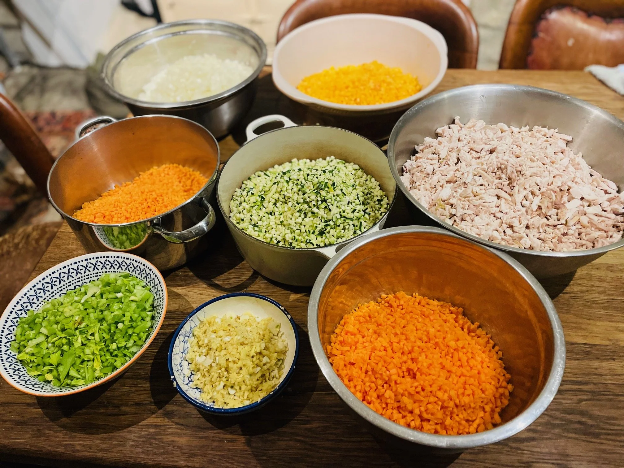 Various bowls filled with chopped vegetables and shredded meat on a wooden table. Vegetables include carrots, zucchini, celery, garlic, and onions.