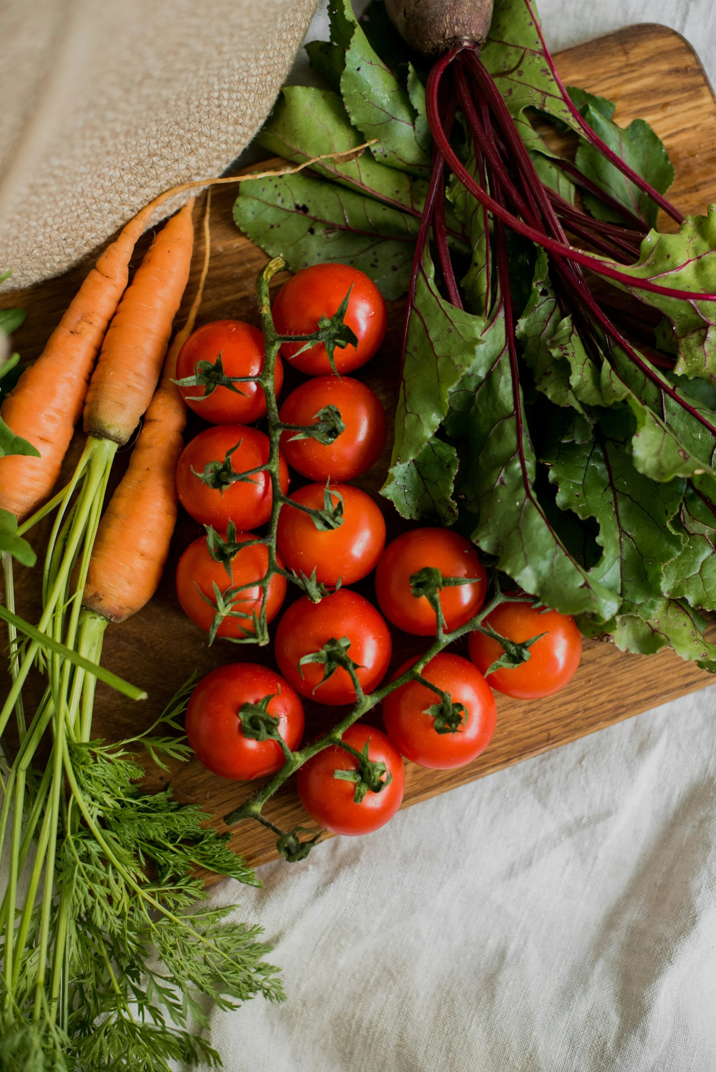 Fresh carrots, cherry tomatoes on a vine, and beet greens on a wooden surface.