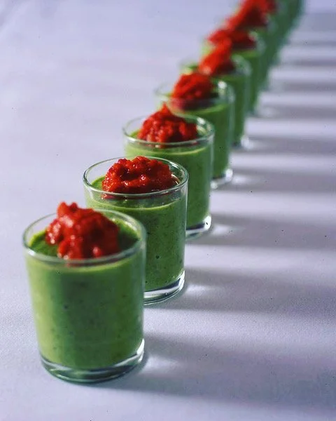 Row of small glasses filled with green soup topped with red sauce on a white surface.