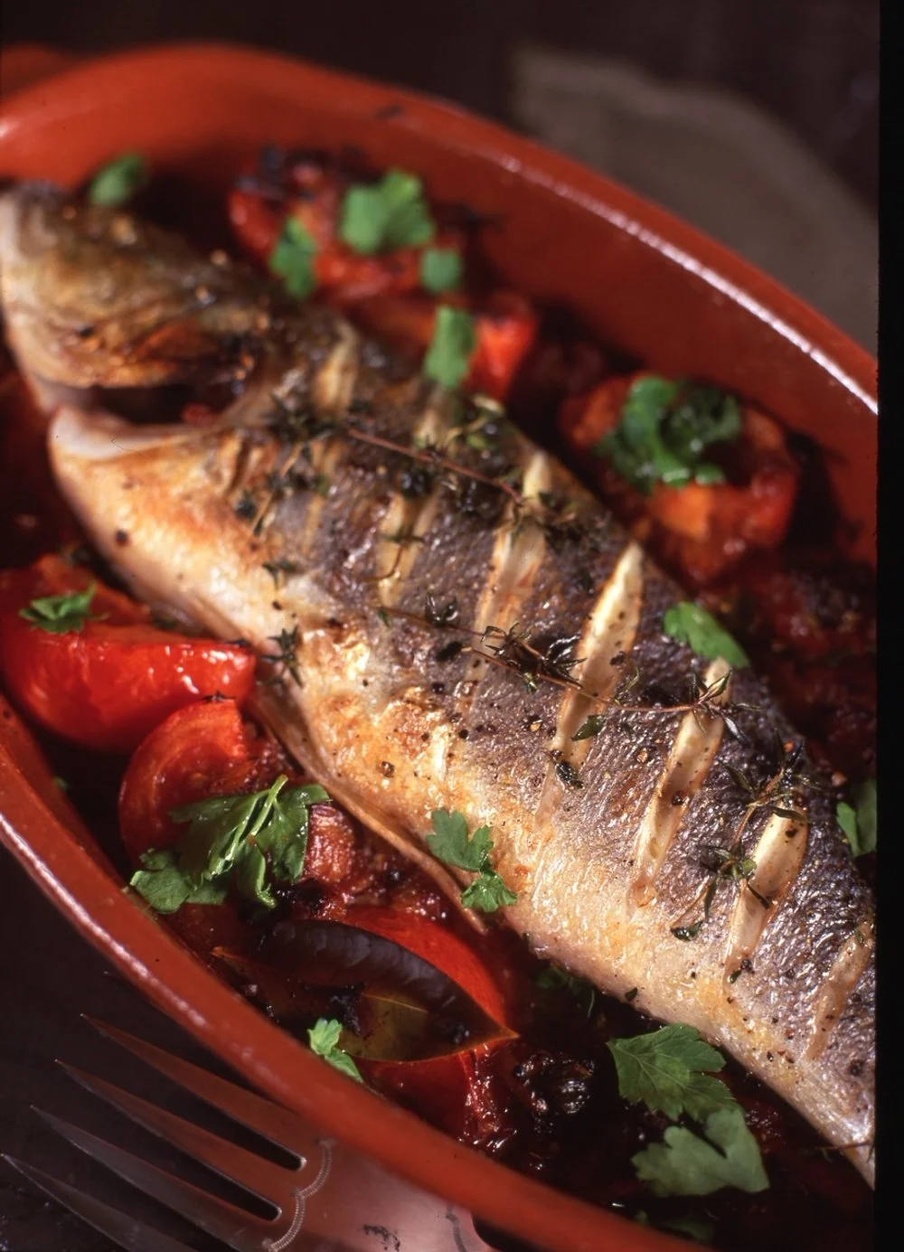 Baked whole fish with herbs, tomatoes, and garnished with parsley in a terracotta dish.