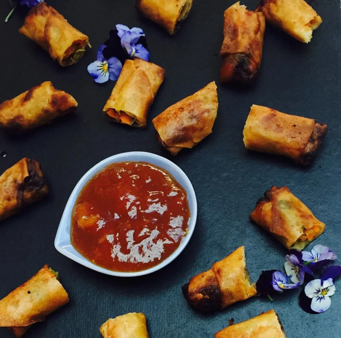 Spring rolls and dipping sauce with edible flowers on dark surface.