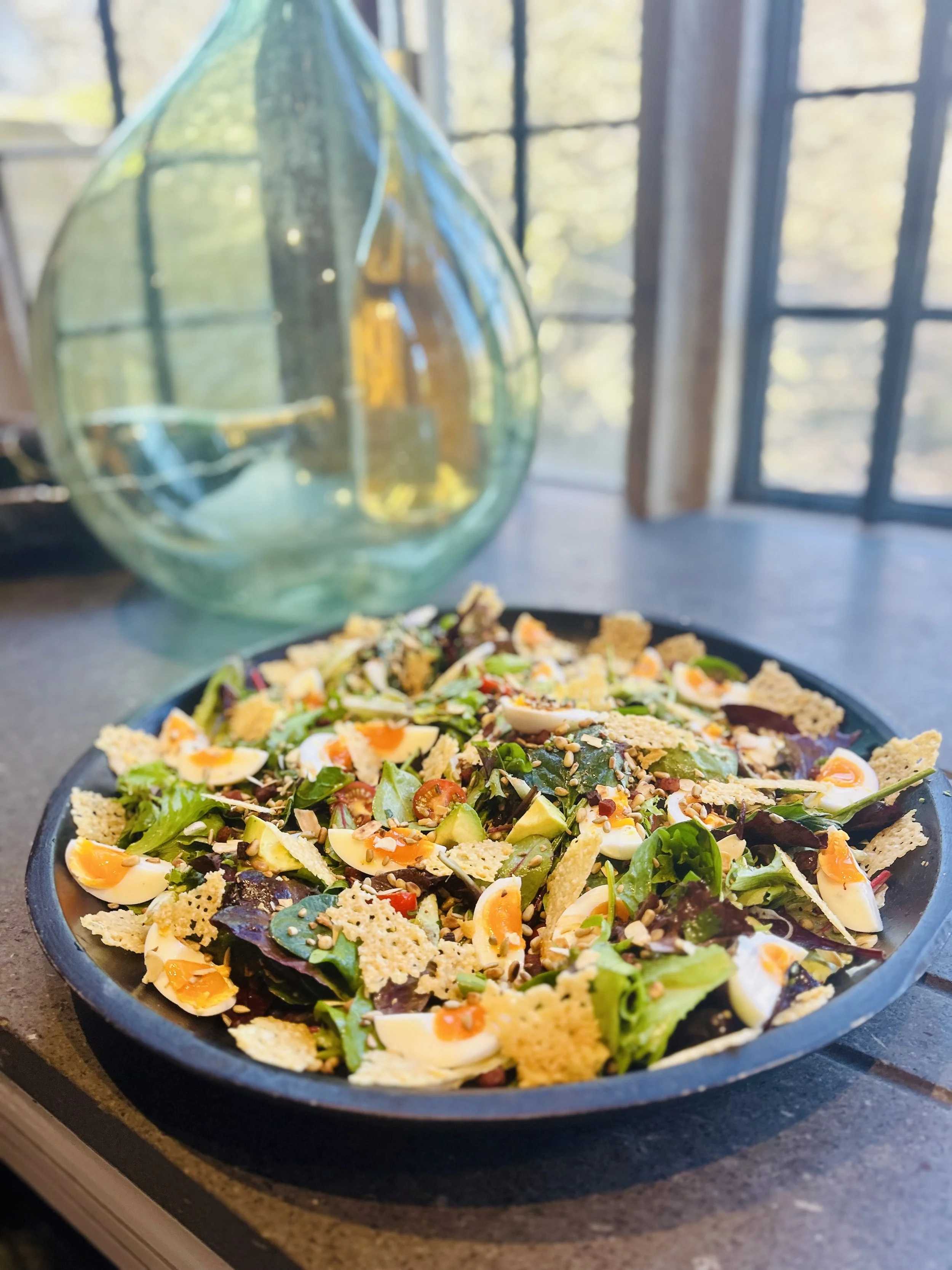 A fresh salad with mixed greens, avocado, boiled eggs, seeds, and cheese crisps on a black plate, placed next to a large glass vase in front of a sunlit window.