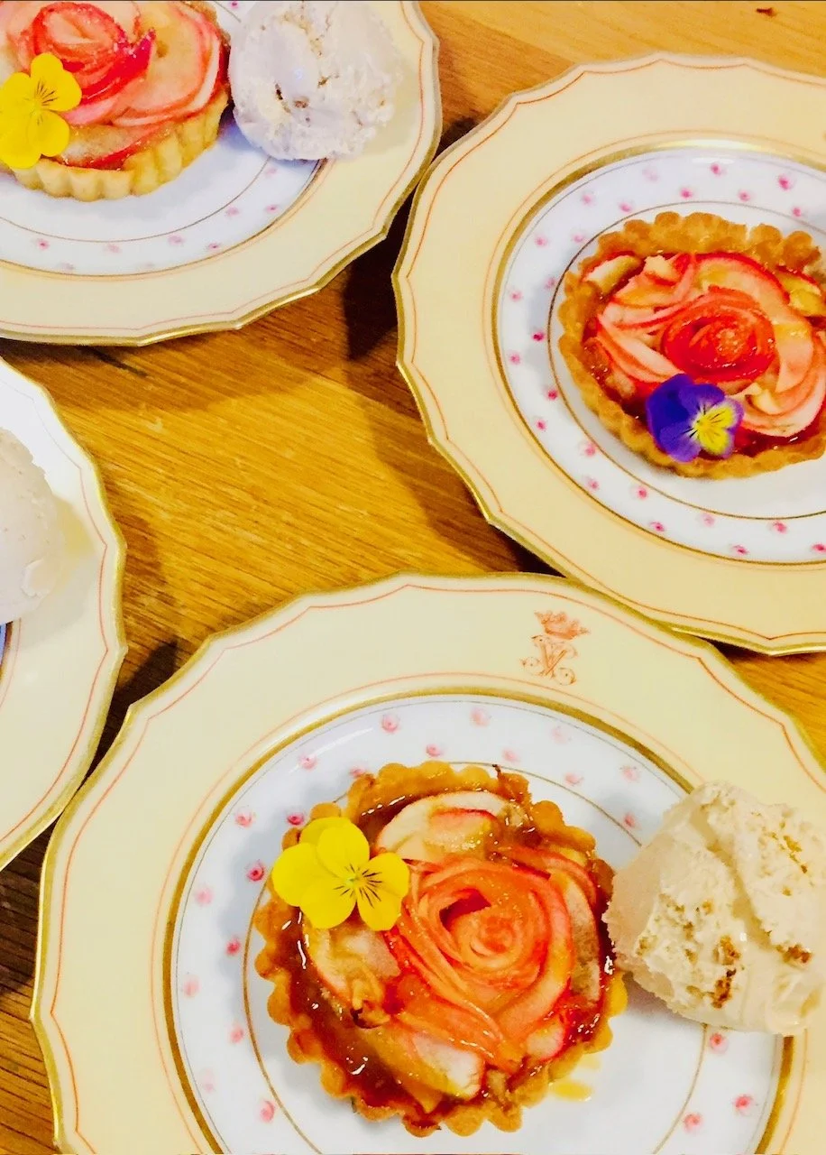 Dessert plates with apple rose tarts, ice cream scoops, and edible flowers on a wooden table.