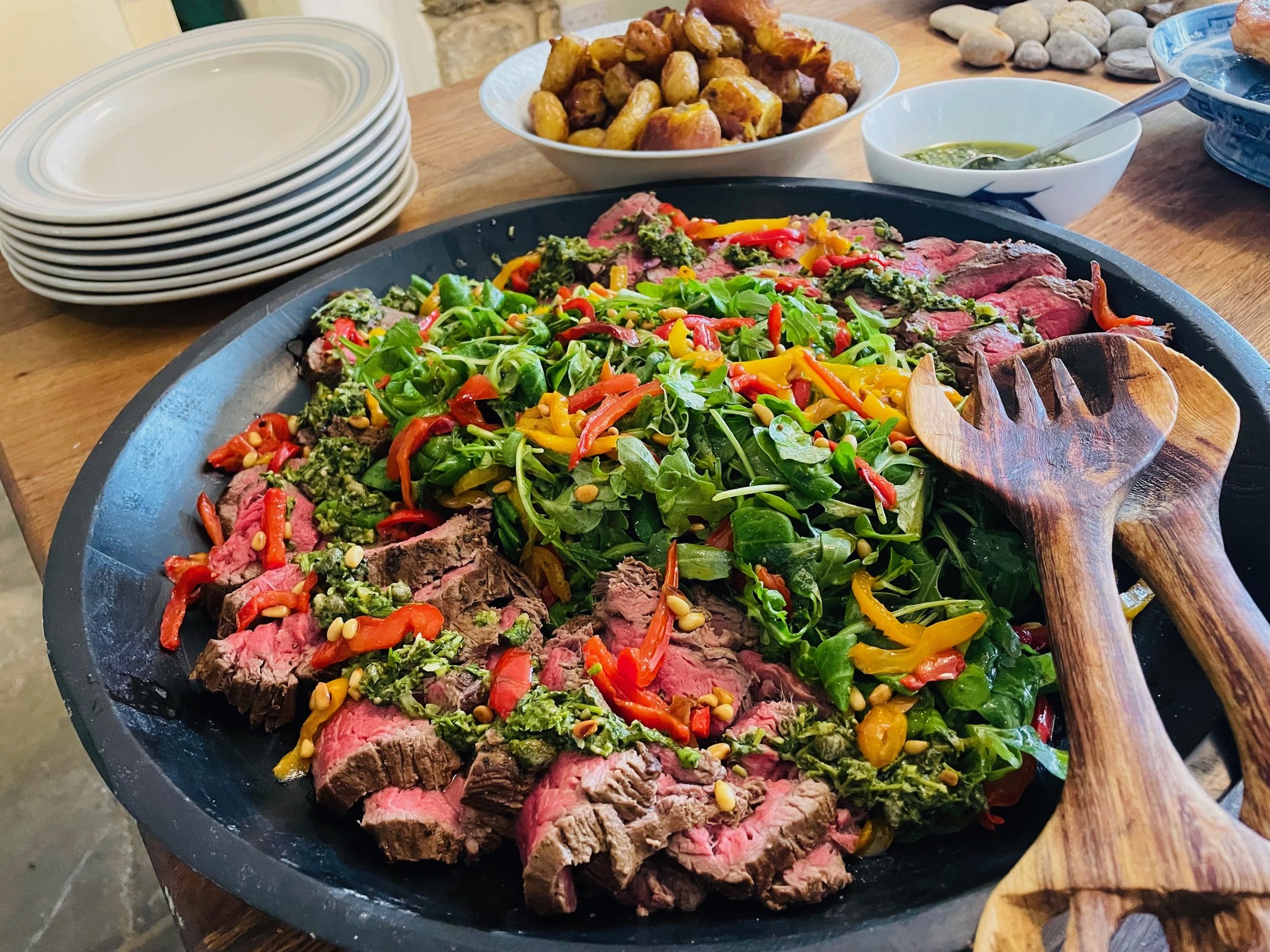 Sliced beef steak on a platter with arugula, roasted red and yellow peppers, pine nuts, and pesto, accompanied by serving spoons; background includes stacked plates and a bowl of roasted potatoes.
