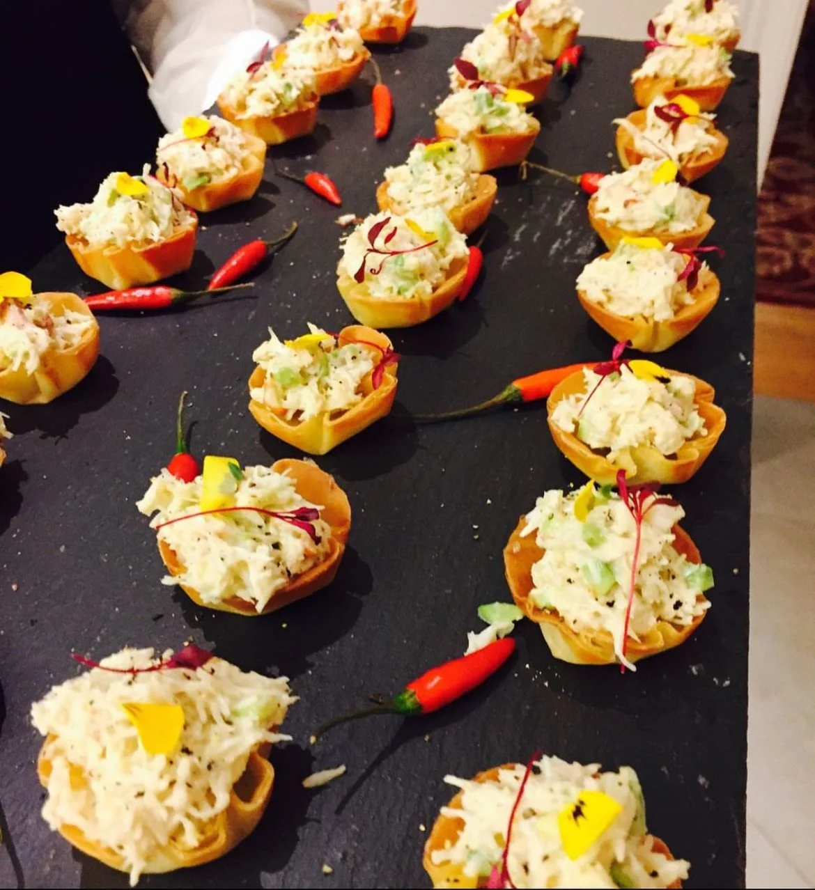 Appetizers on a slate platter, featuring crisp pastry cups filled with a creamy salad mixture, garnished with microgreens and yellow edible flowers, alongside small red chili peppers.