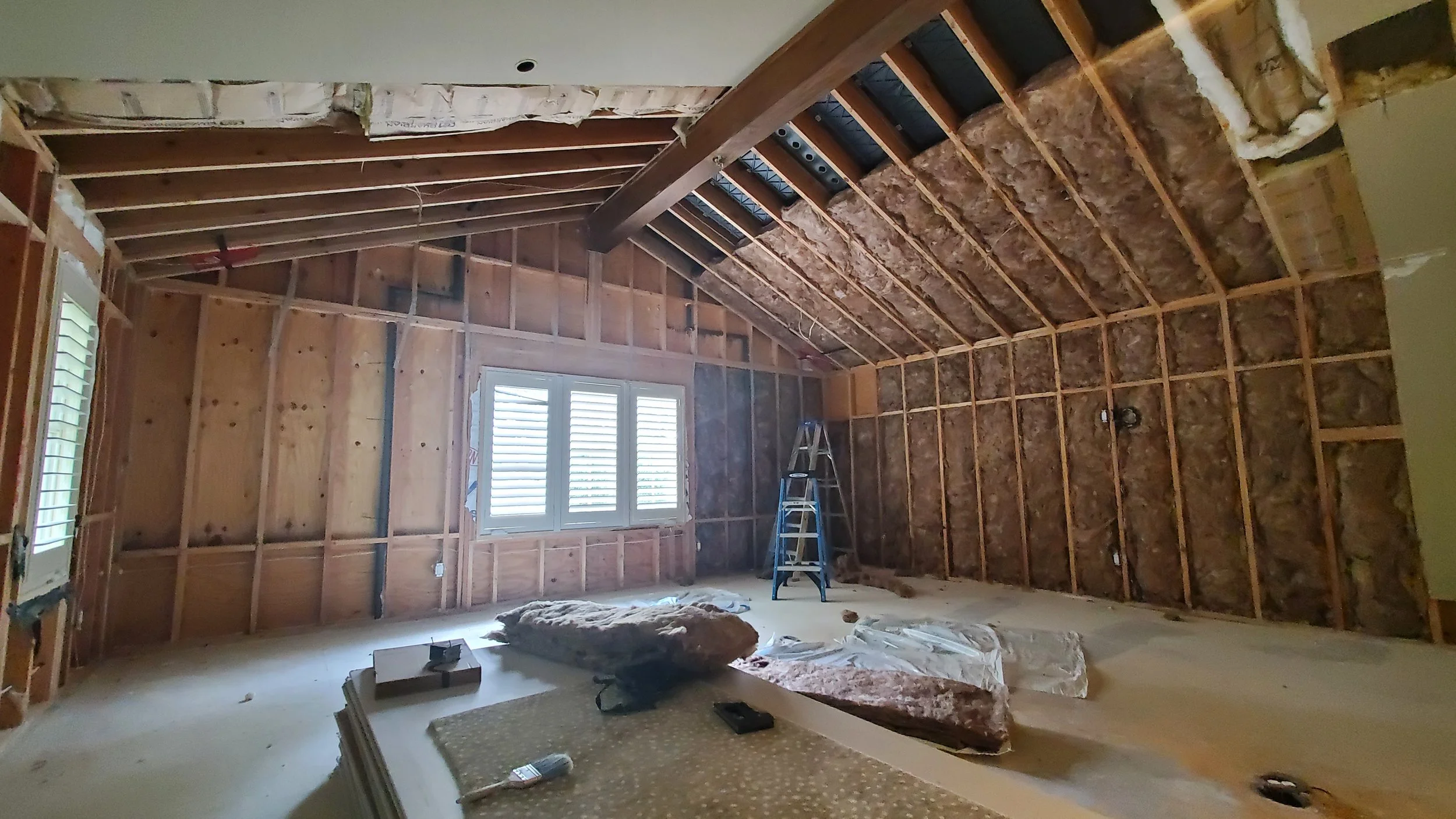 Open room with a sloped ceiling, exposed wall framing and insulation, a shuttered window, and a ladder, before drywall in a Memorial, Houston remodel