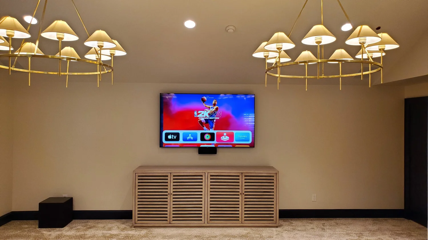 Finished media room with a wall-mounted TV on a louvered cabinet between two chandeliers